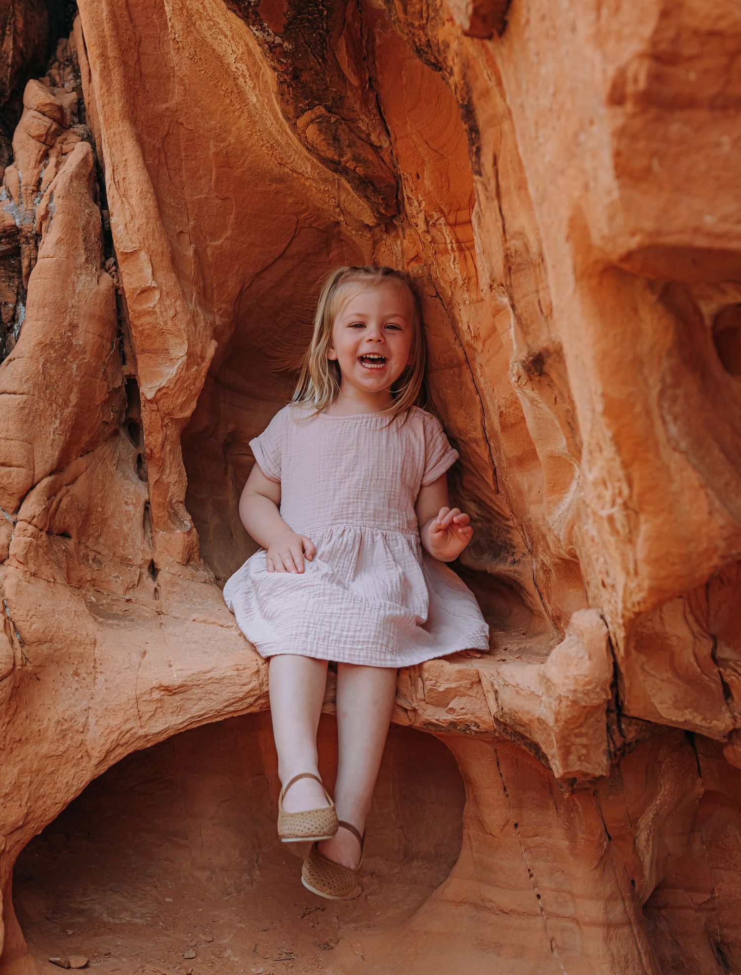 Laughing young girl in pink dress sitting in a natural red sandstone rock alcove.