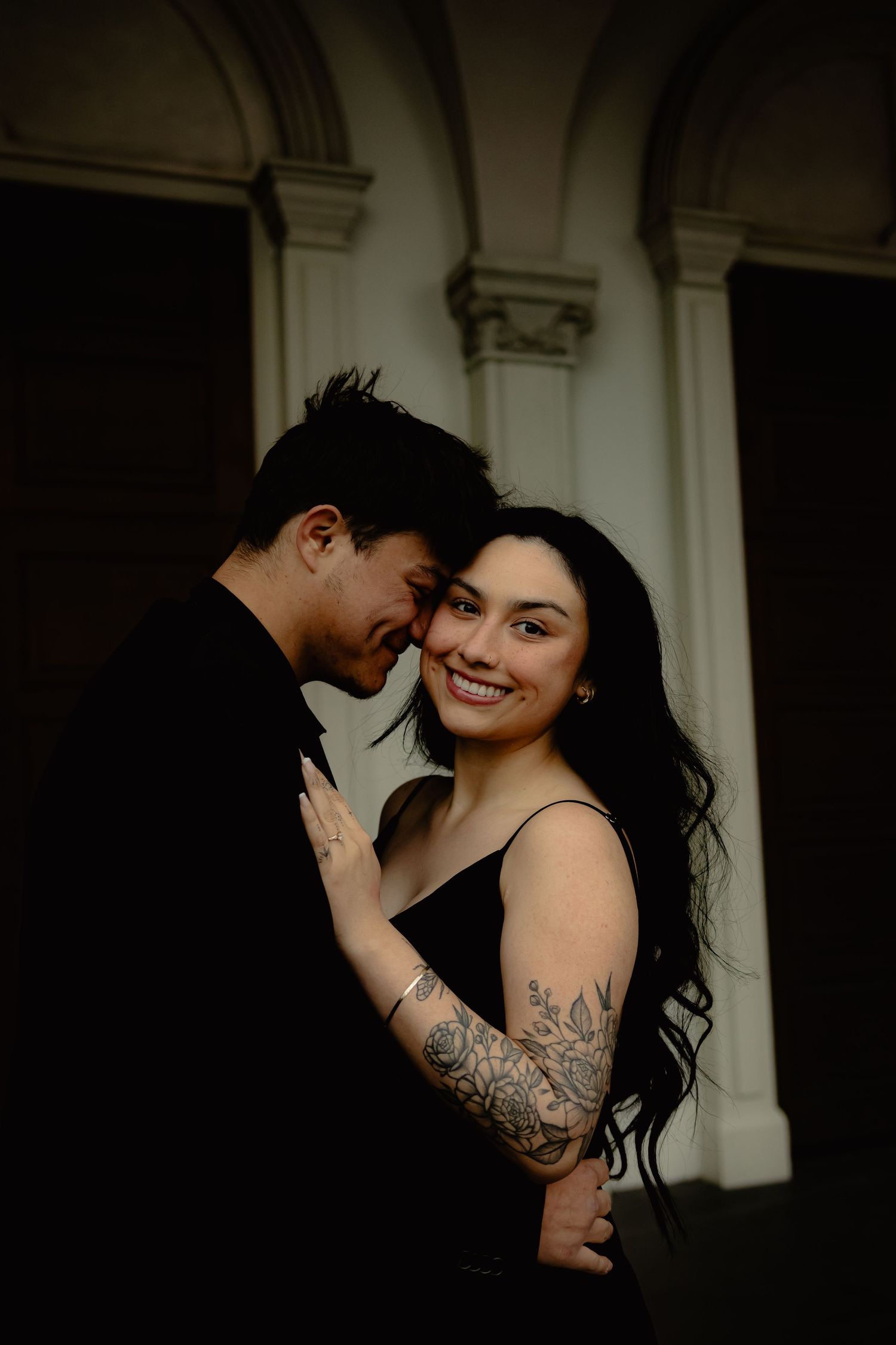 Smiling woman with floral arm tattoo in black dress embracing partner during romantic engagement shoot.