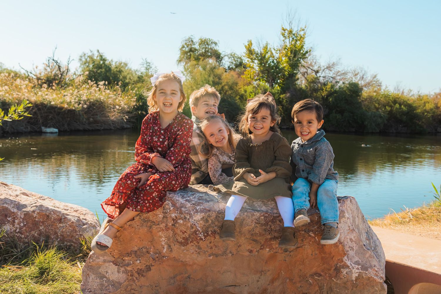 Five children sitting on a rock by a pond in a sunny outdoor setting.