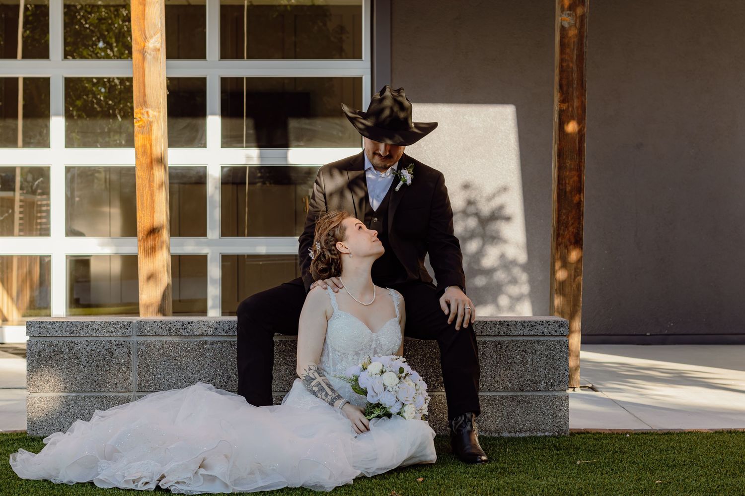 Bride sits on stone bench leaning against groom in cowboy hat holding white bouquet outdoors.