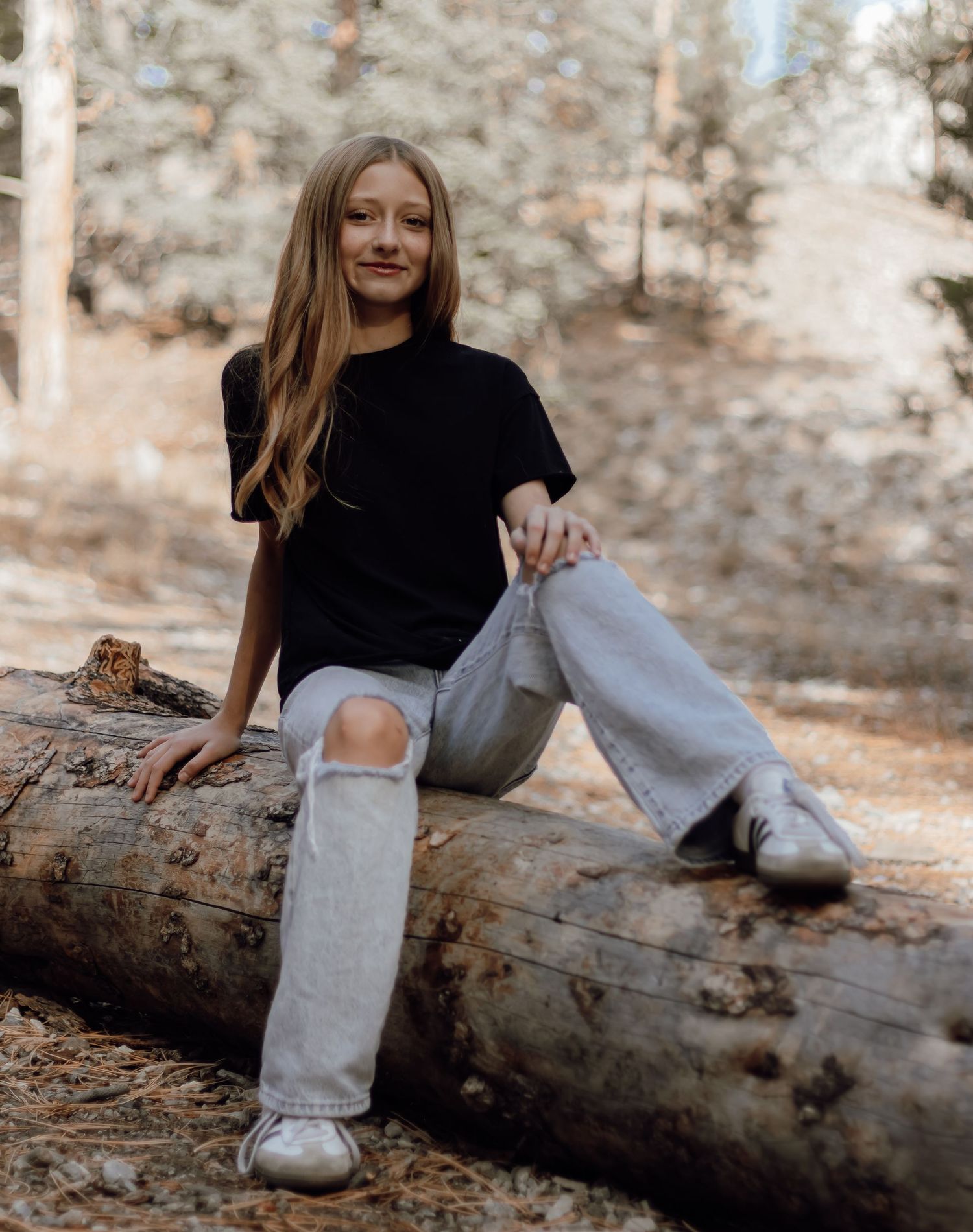 A teenage girl in a black tee and ripped jeans sits on a log in a sunlit forest.