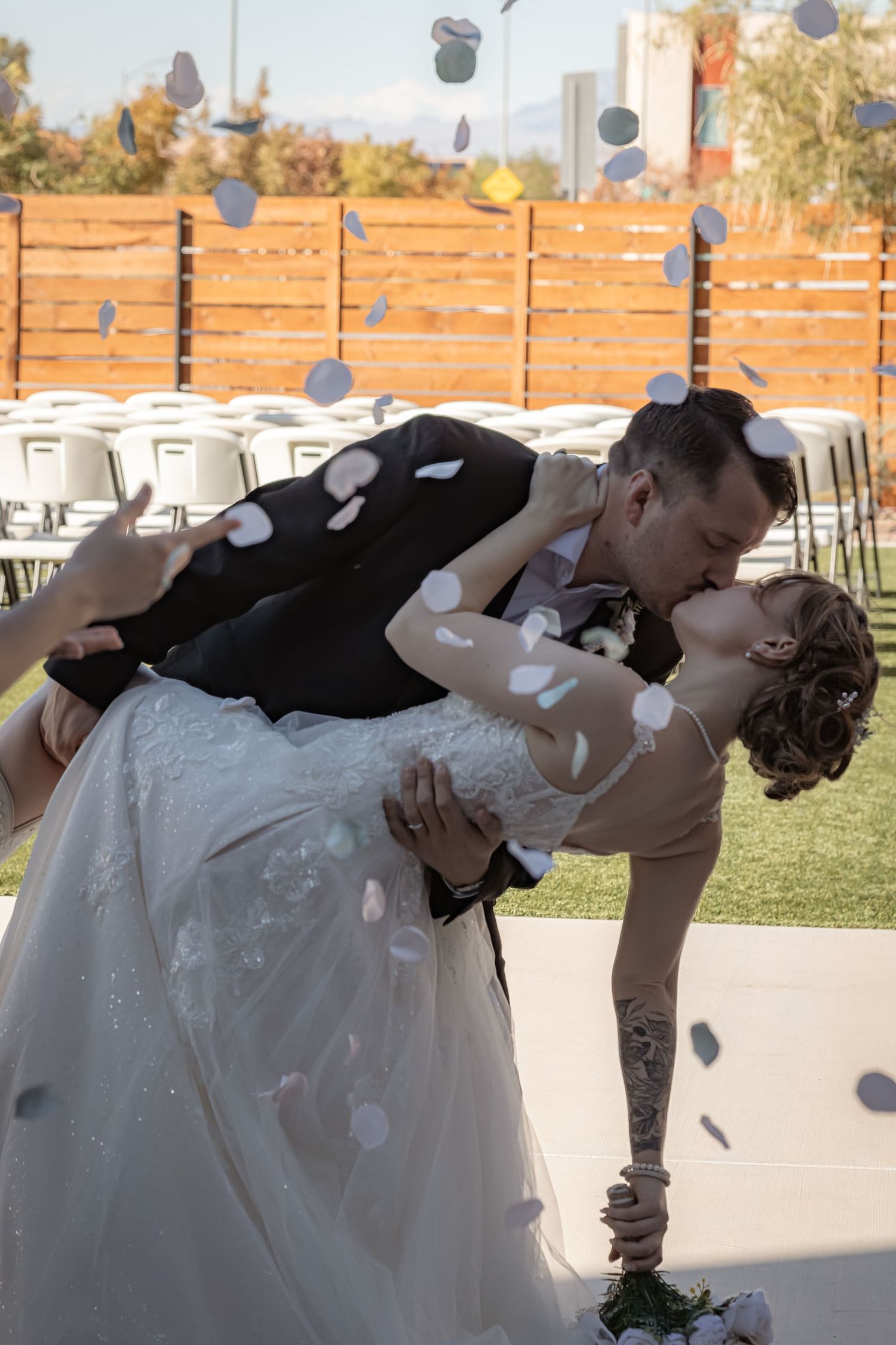 Groom dips bride for a kiss as confetti falls during outdoor wedding ceremony celebration.