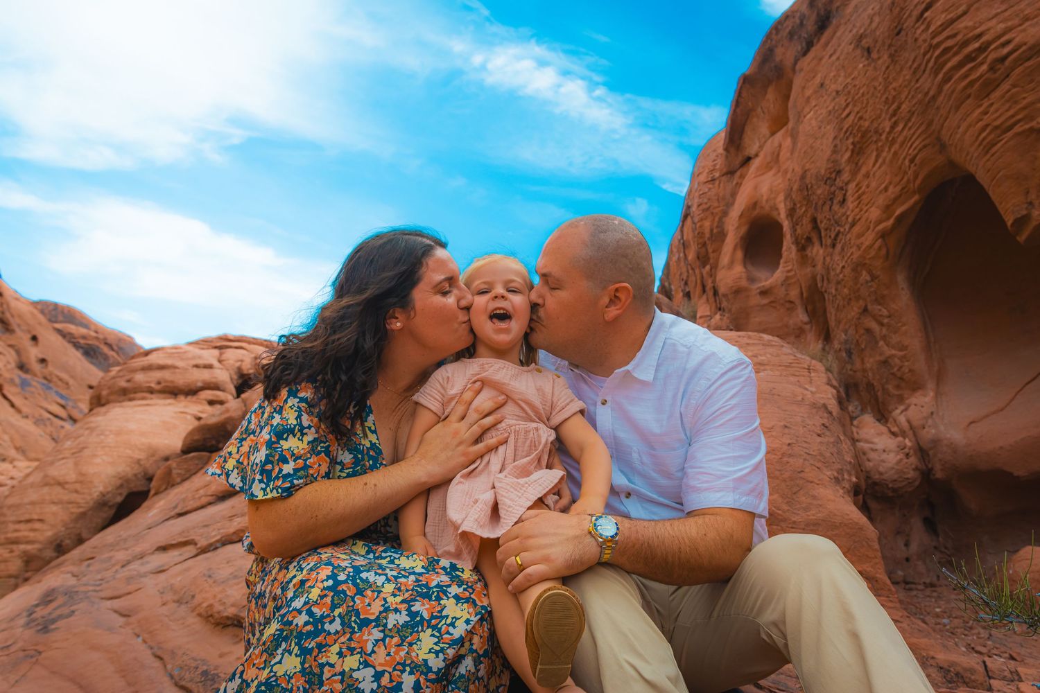 Parents kissing their young daughter on the cheeks while sitting on red rocks outdoors.