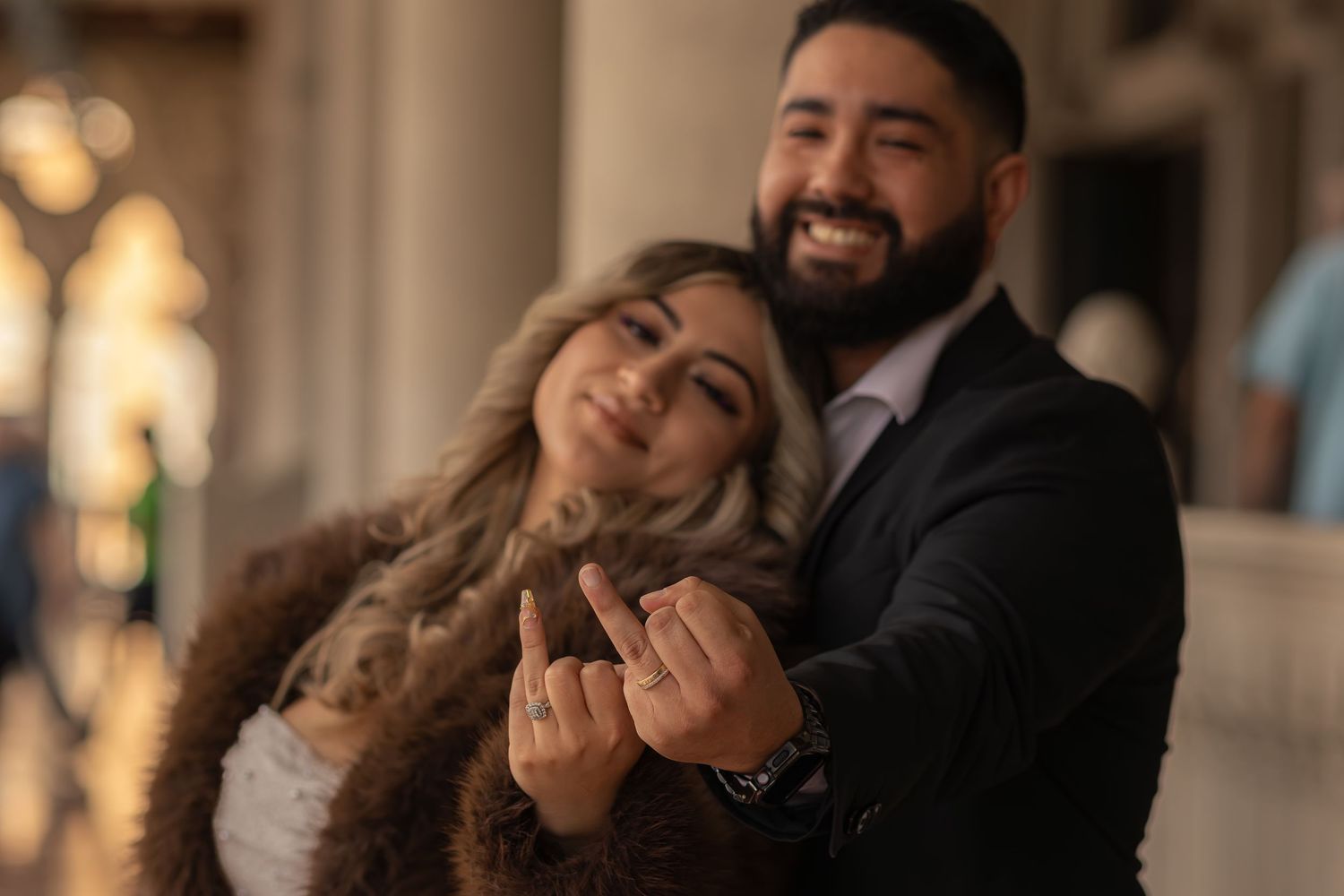 Smiling couple showing off engagement ring at a warmly lit venue.