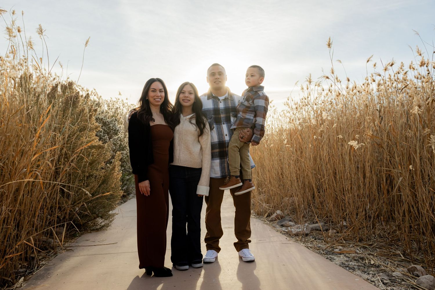 Family of four posing on a sunlit path surrounded by tall golden reeds in a nature setting.
