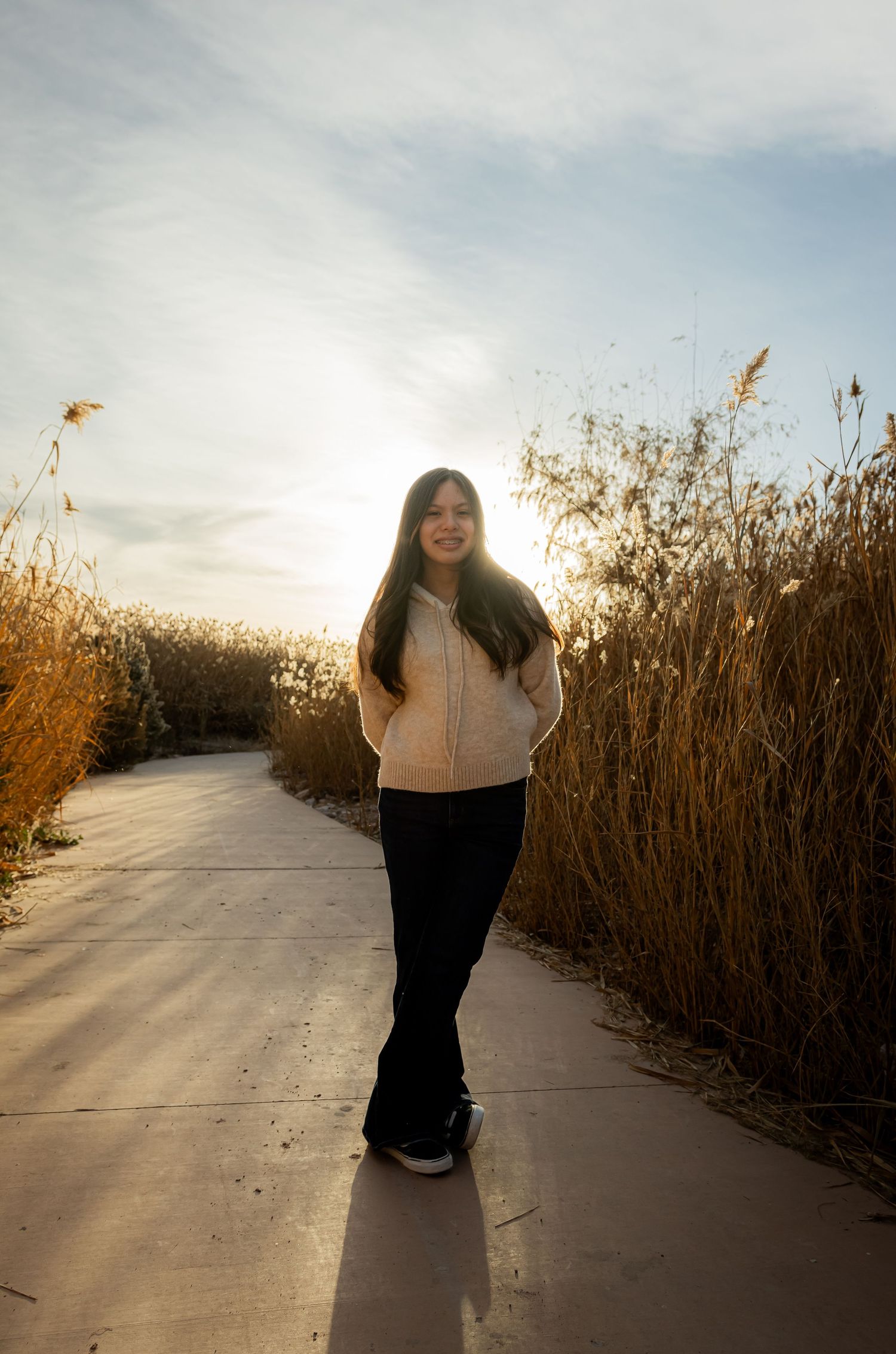 Smiling young woman in a cream sweater and flare jeans standing on a sunlit path among tall golden grass.