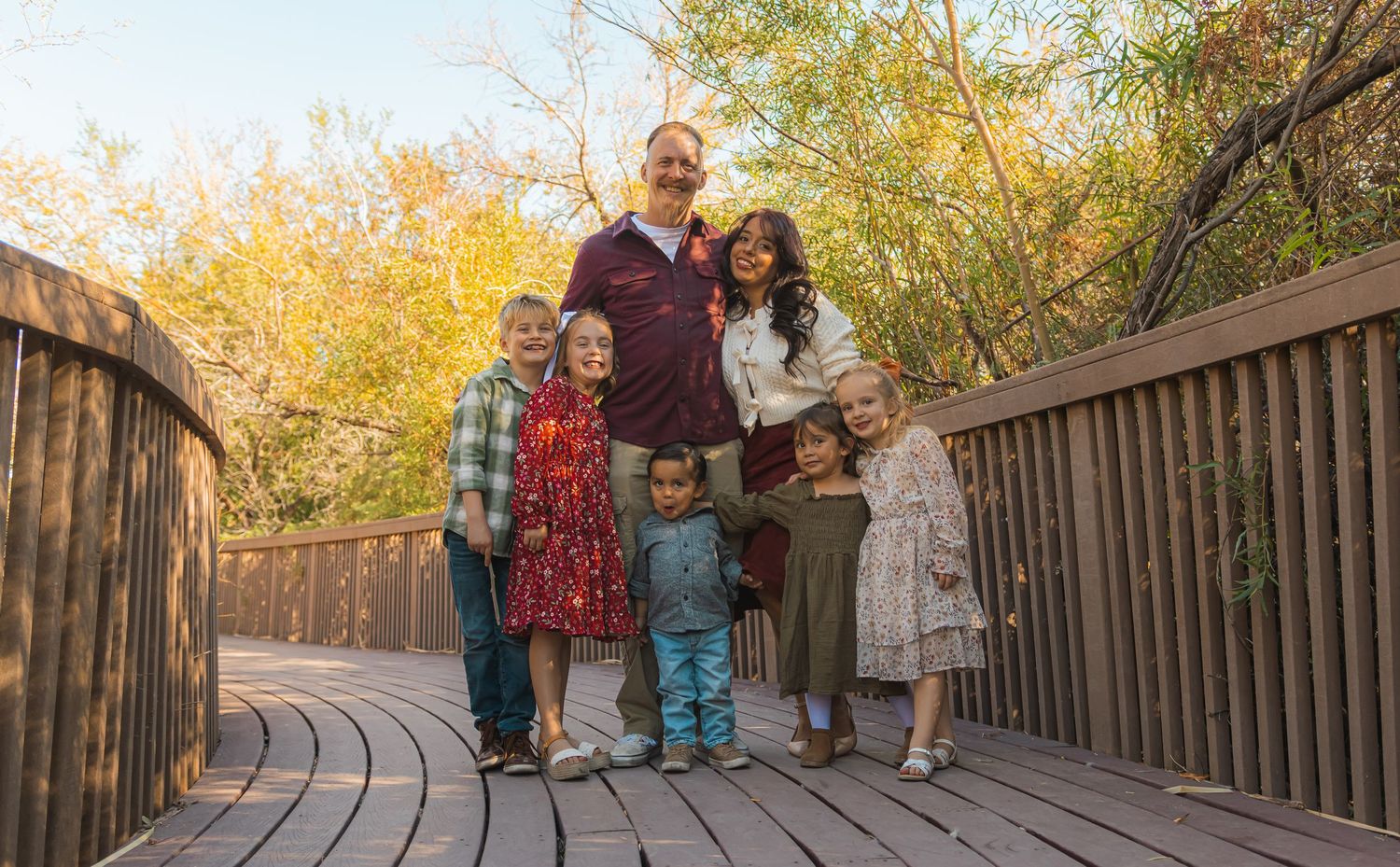 Family of seven smiling on a wooden bridge surrounded by autumn trees.