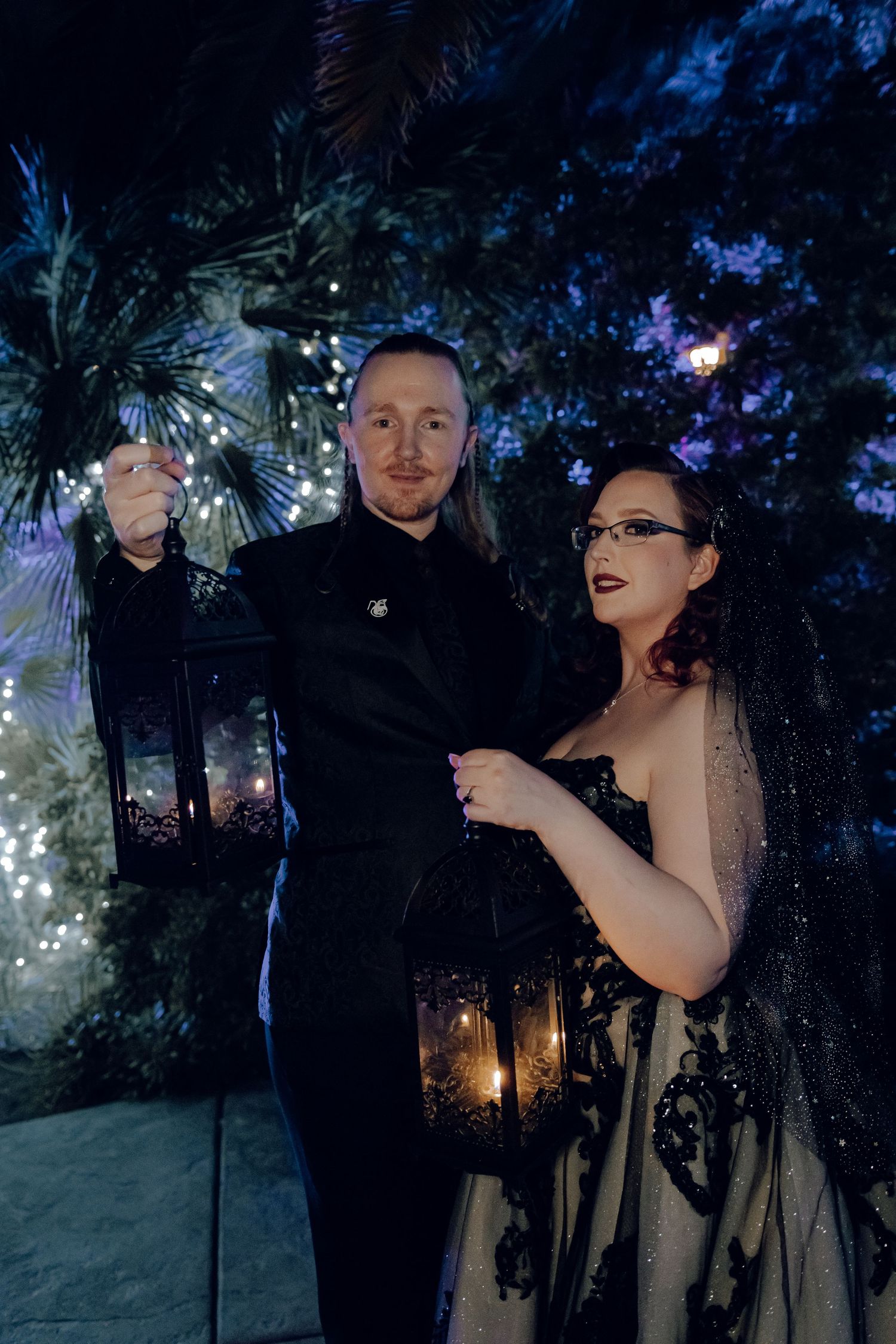 Gothic wedding couple holding ornate black lanterns at night amid twinkling lights and dark foliage.