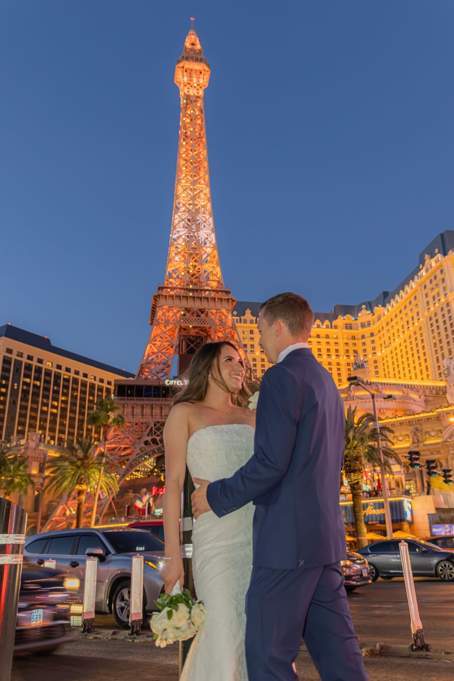 Bride and groom pose romantically in front of the illuminated Eiffel Tower replica at Paris Las Vegas at dusk.