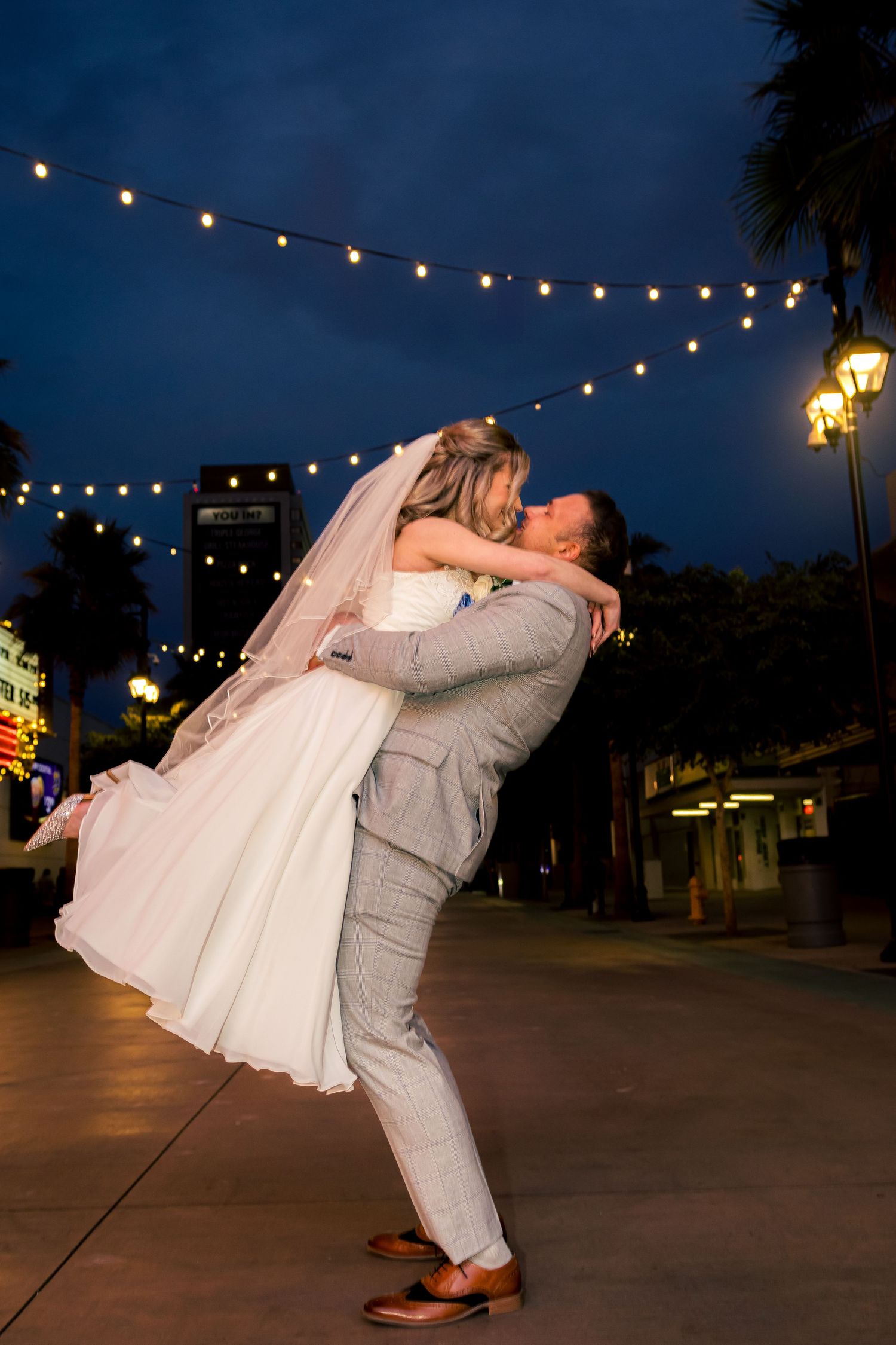 Groom lifting bride in white gown for romantic kiss under string lights at night in Las Vegas.