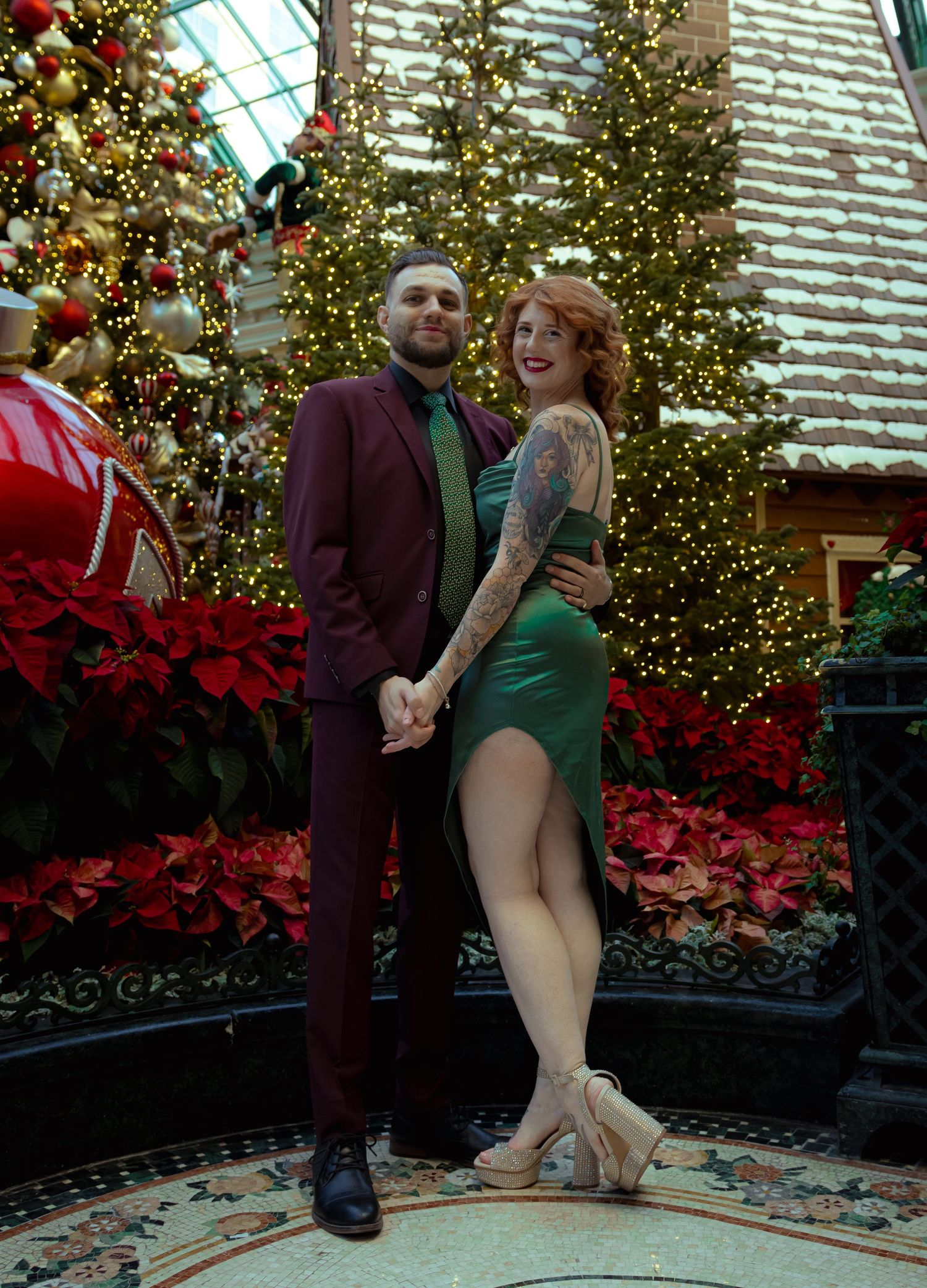 Couple posing in front of Christmas trees and poinsettias, man in burgundy suit, woman in green dress with tattoos.
