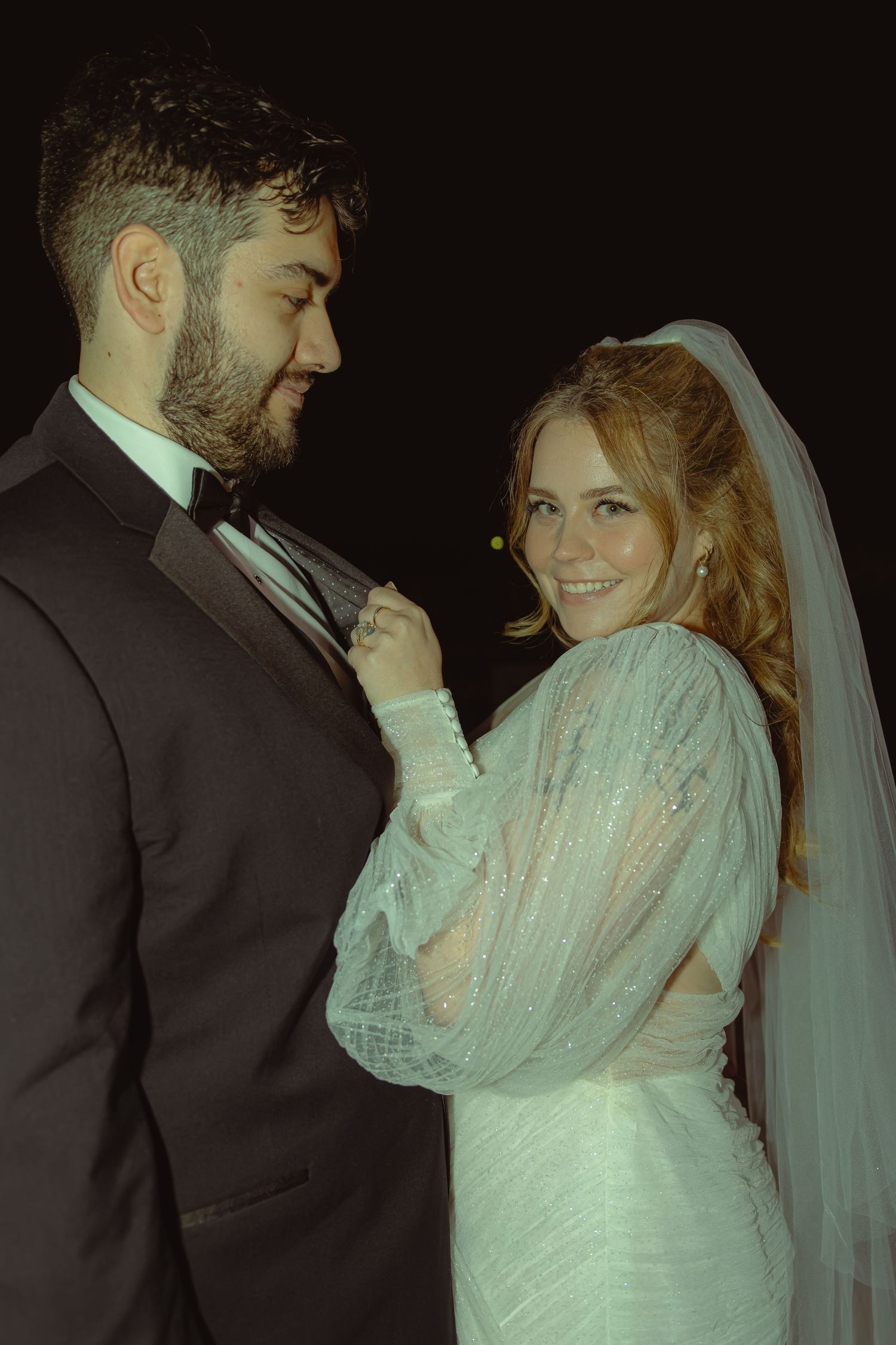 A smiling bride in a white lace gown and veil adjusts her groom's bow tie as they pose together at their wedding.