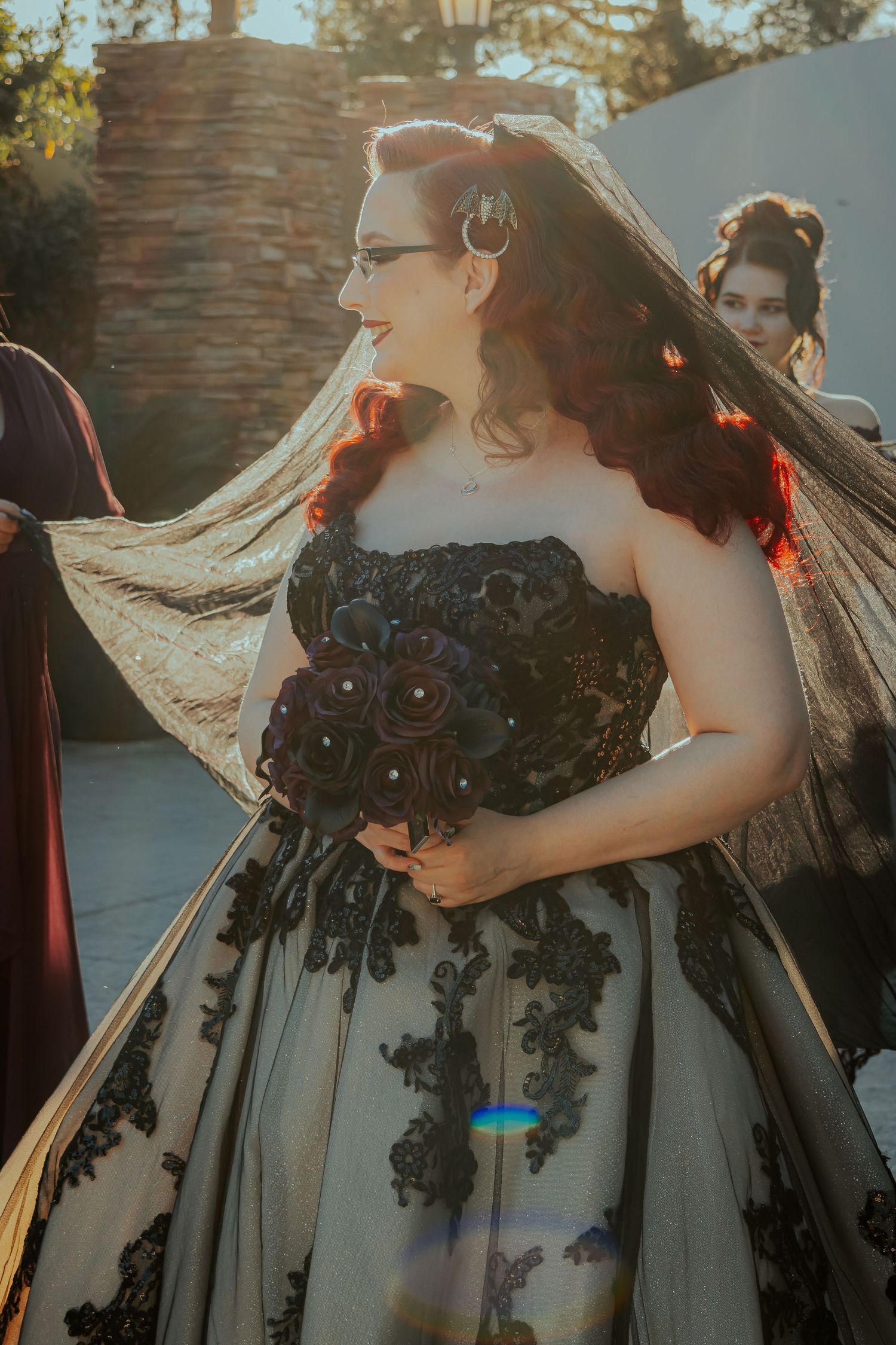 Gothic bride in black lace strapless gown with flowing veil holding dark floral bouquet outdoors.