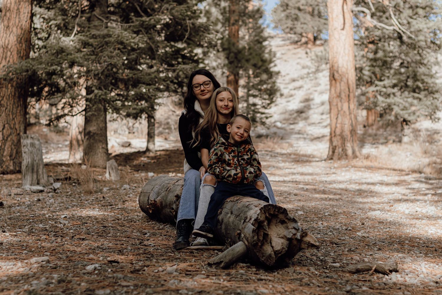 Woman with two young children sitting on a fallen log in a pine forest setting.