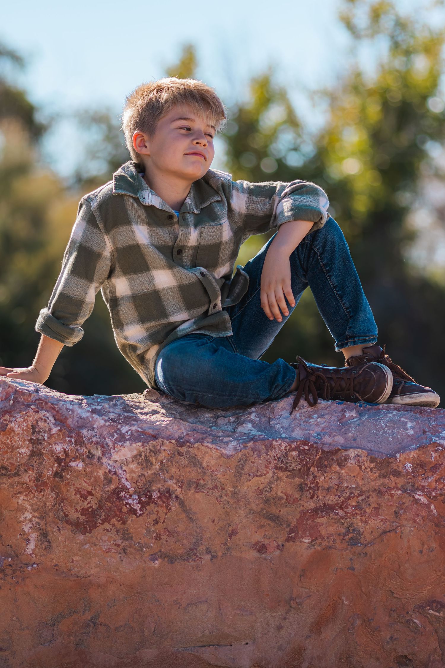 Boy in a plaid hoodie sitting relaxed on a large rock outdoors.