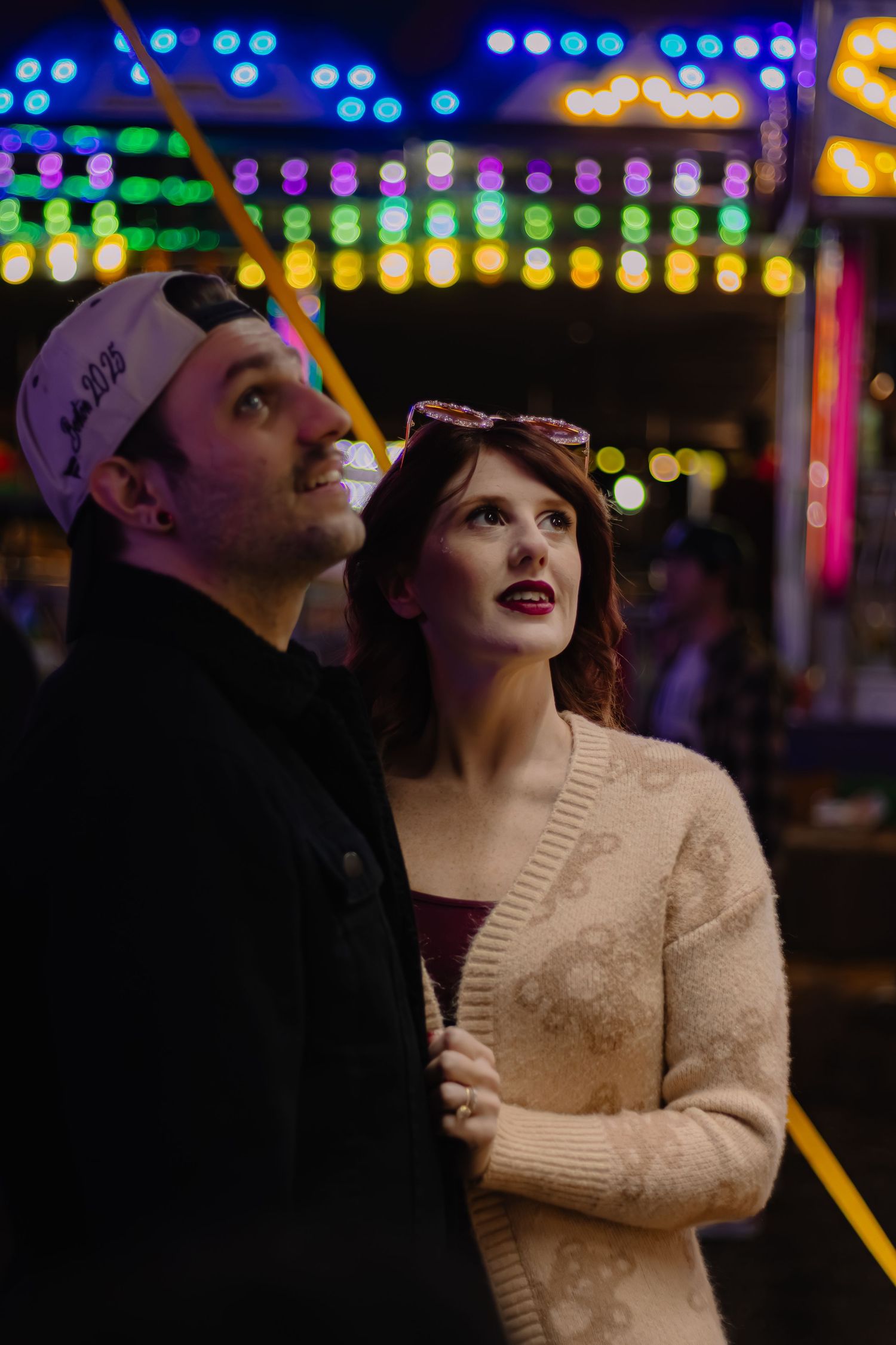Couple gazing up at colorful carnival lights at night, woman in cream sweater with red lips, man in baseball cap.