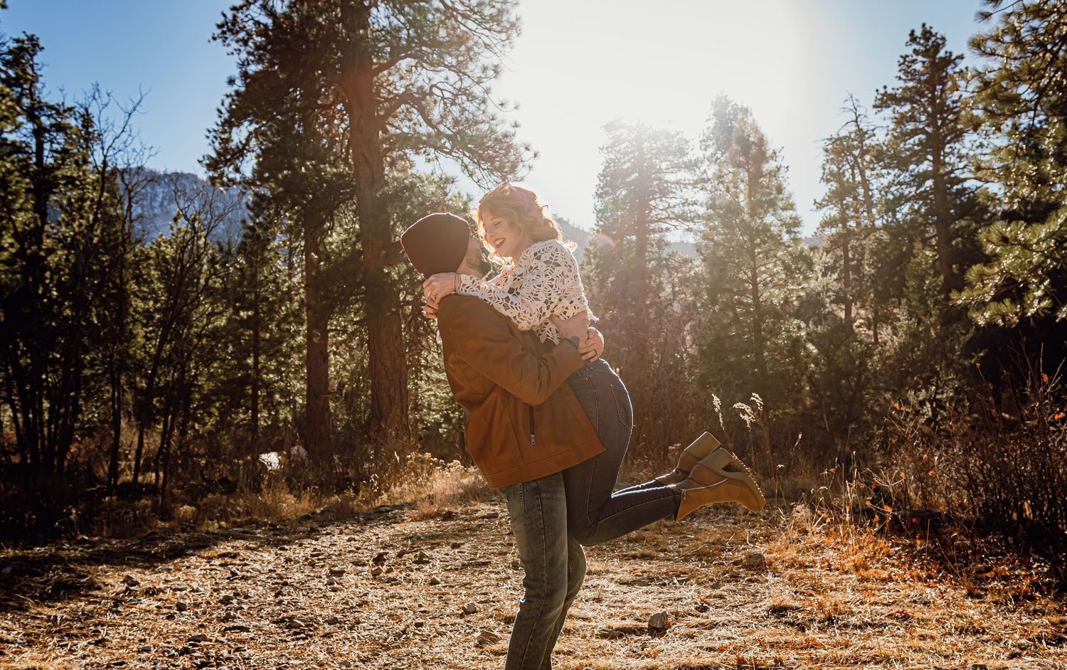 Couple embracing in sunlit forest clearing, man lifting woman romantically among tall pine trees in golden autumn light.