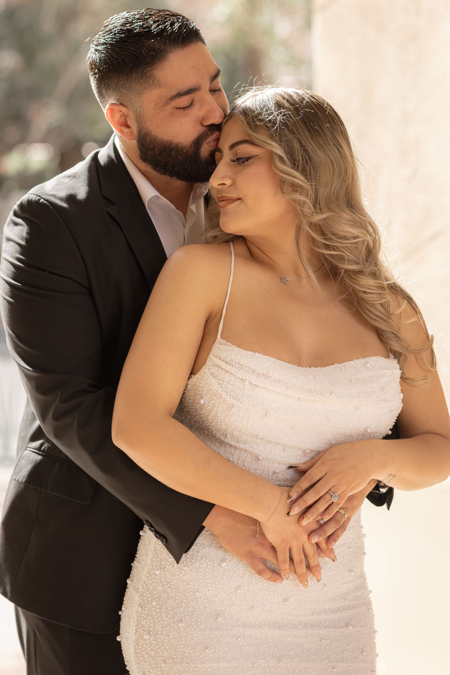 Bride in white beaded gown embraced by groom in dark suit sharing a tender kiss during wedding photos.