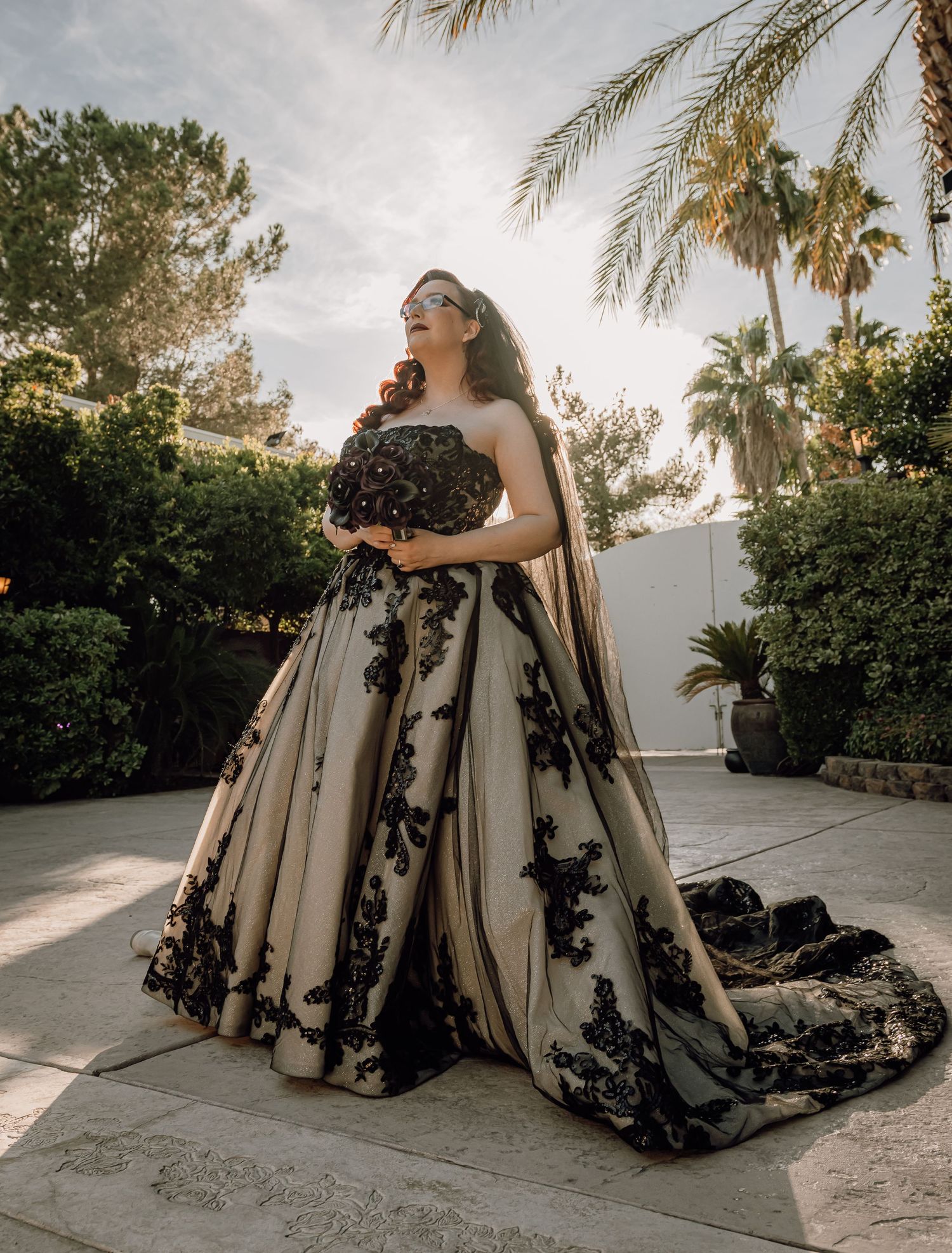 Bride in stunning black lace and champagne ball gown with long train poses in sunlit garden setting.