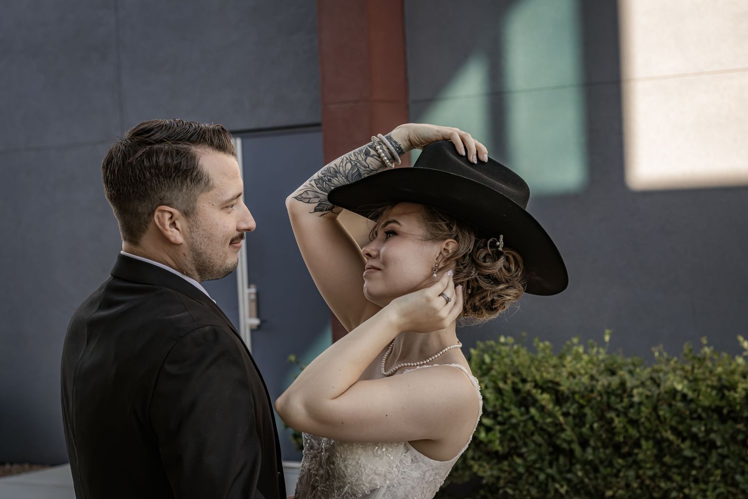 Bride places black cowboy hat on groom's head while smiling outdoors on their wedding day.