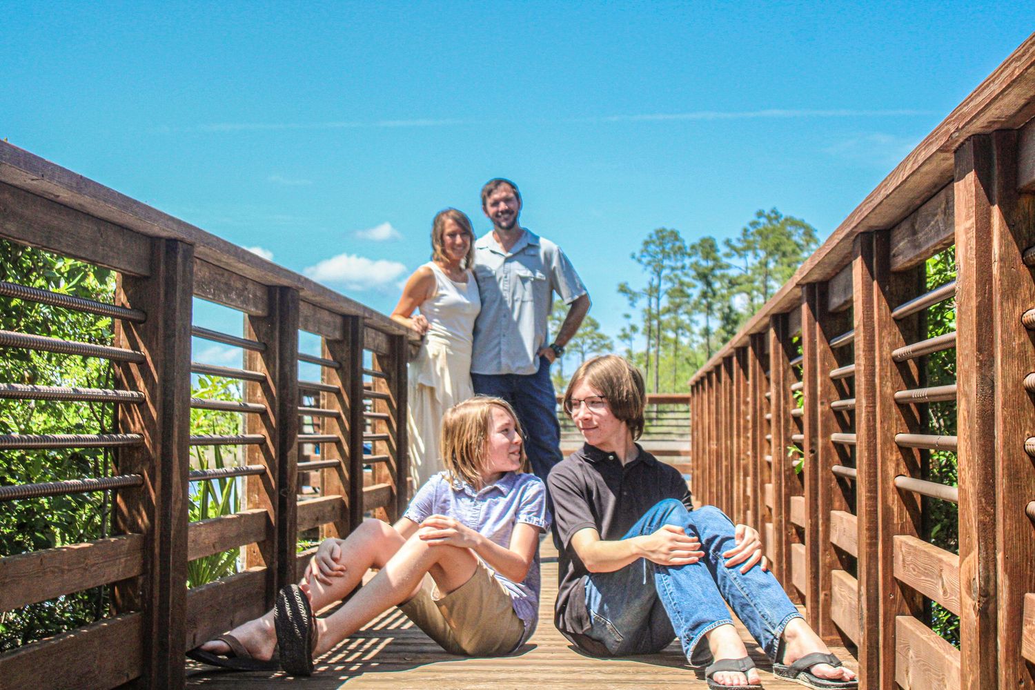Family of four on wooden bridge boardwalk surrounded by lush green nature on sunny day.
