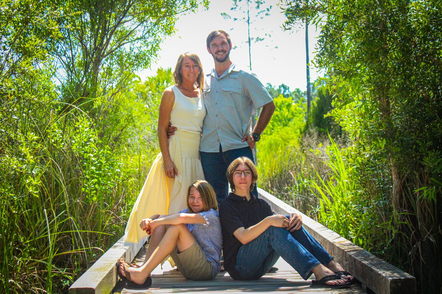 Family of four posing on a wooden bridge surrounded by lush green trees on a sunny day.