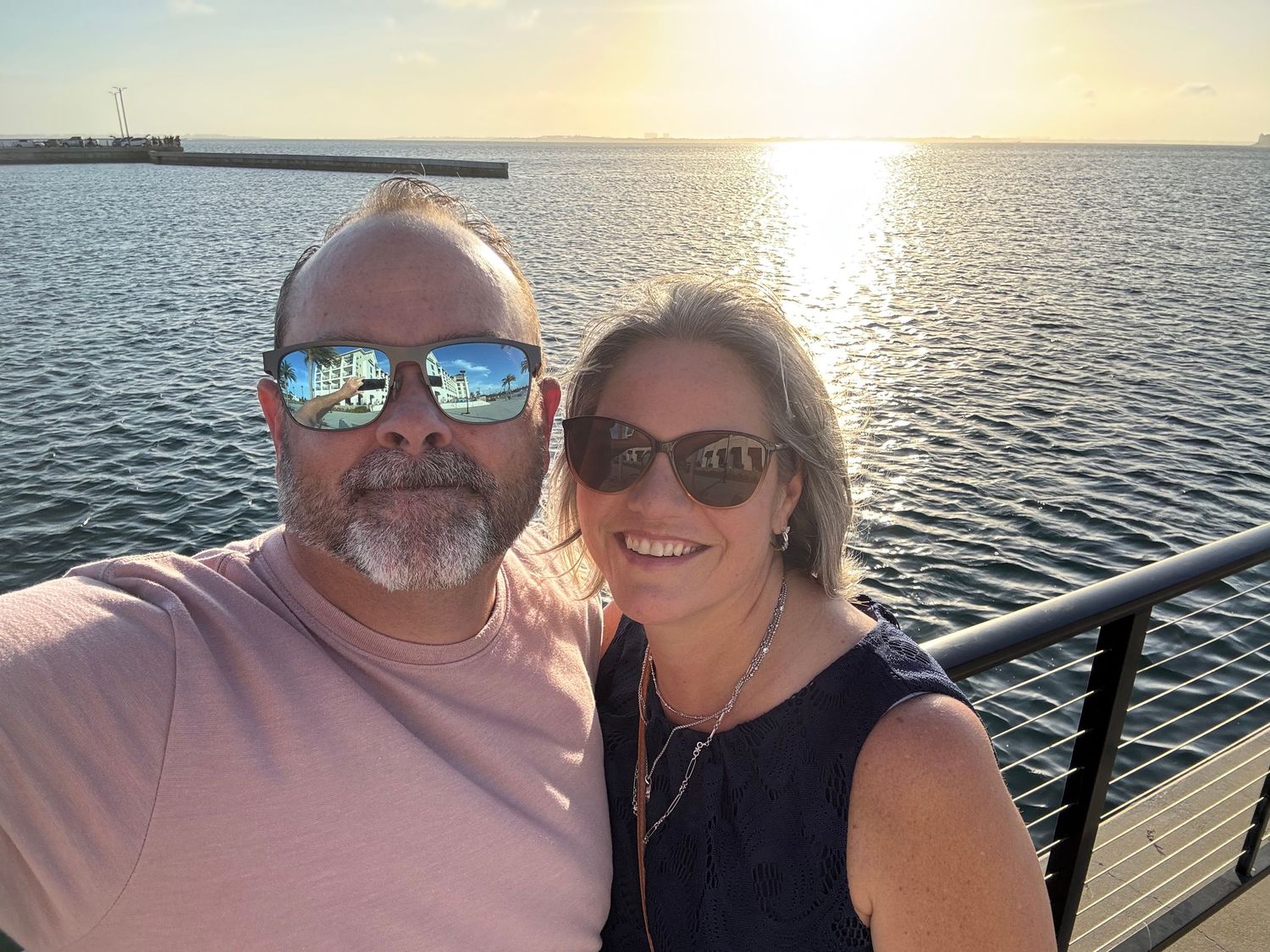 Smiling couple in sunglasses posing for selfie at waterfront during beautiful golden sunset.