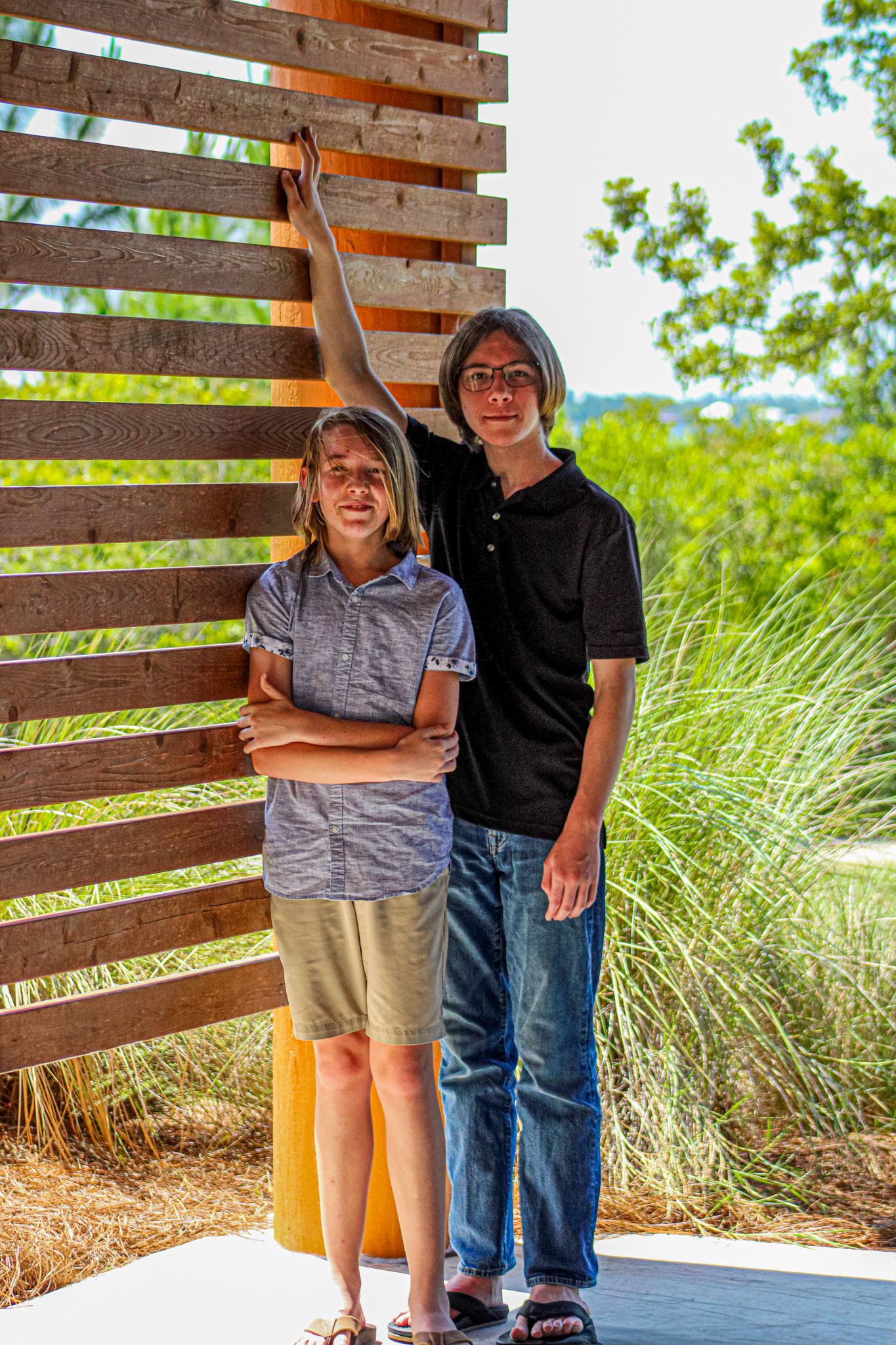 Two young teens with glasses pose beside a wooden slatted wall outdoors, surrounded by lush green trees.