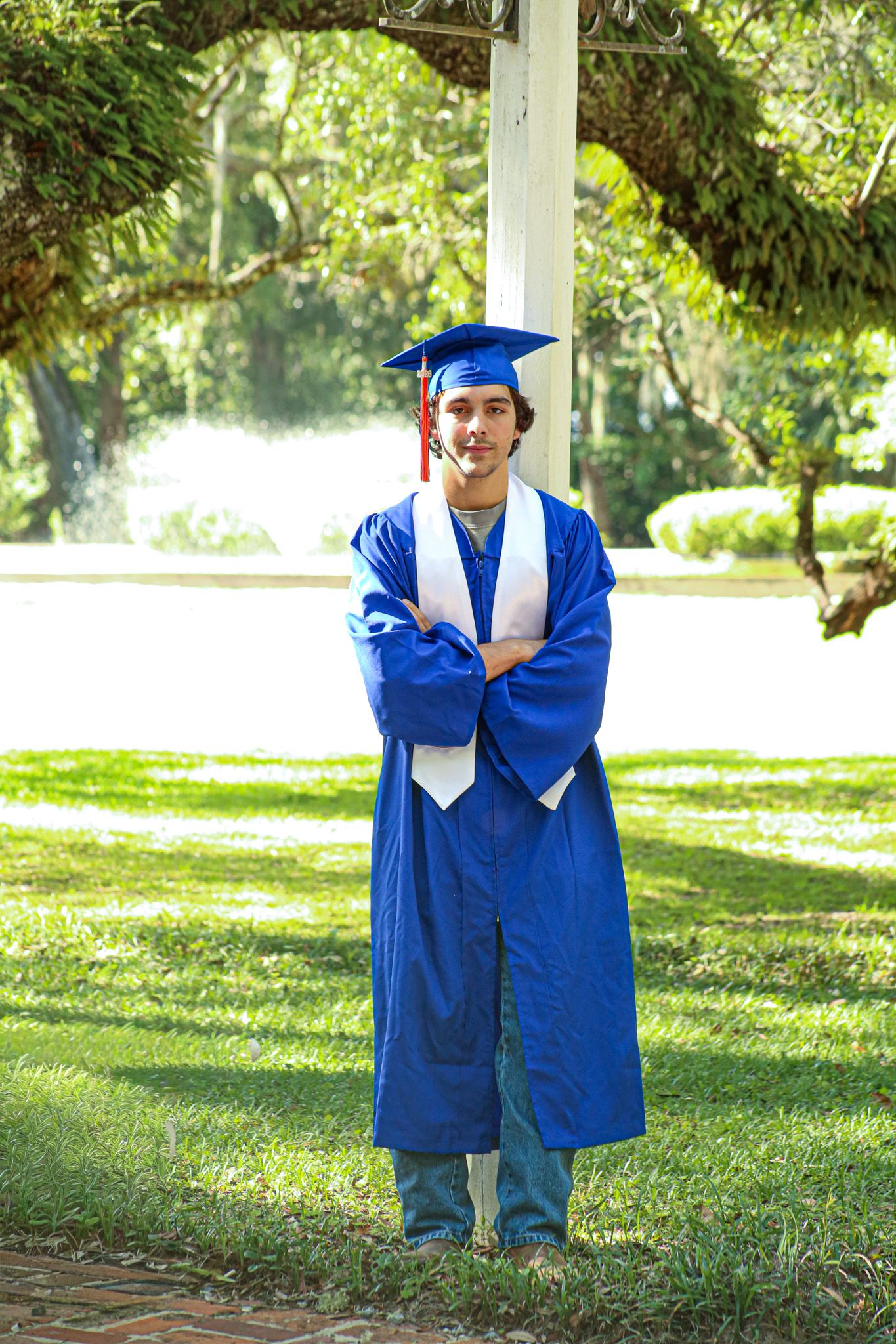 Young man in blue graduation cap and gown stands proudly with arms crossed outdoors in a lush green park. senior year