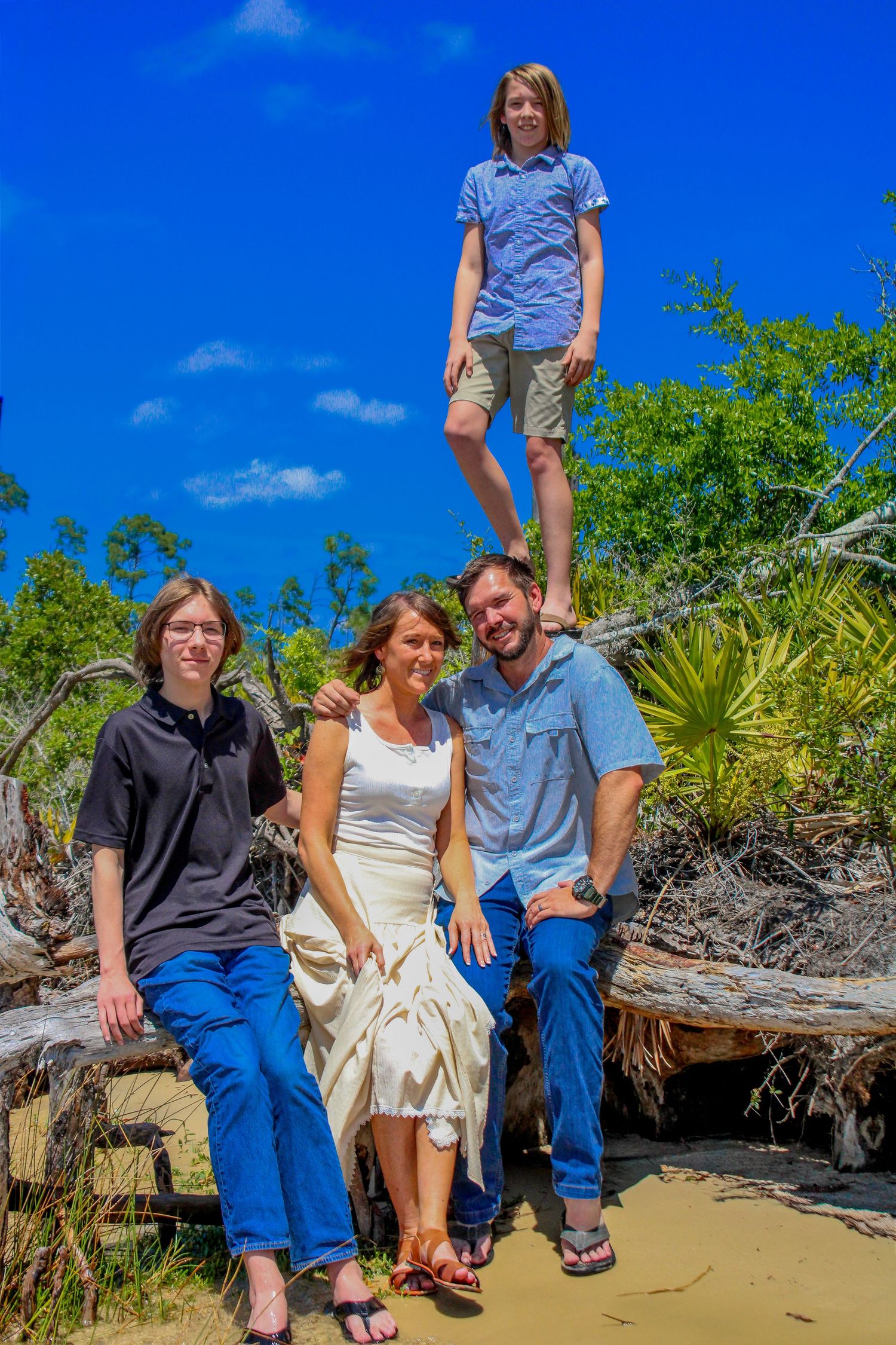 Family of four posing outdoors on driftwood with lush greenery and blue sky, one teen standing elevated above the group.