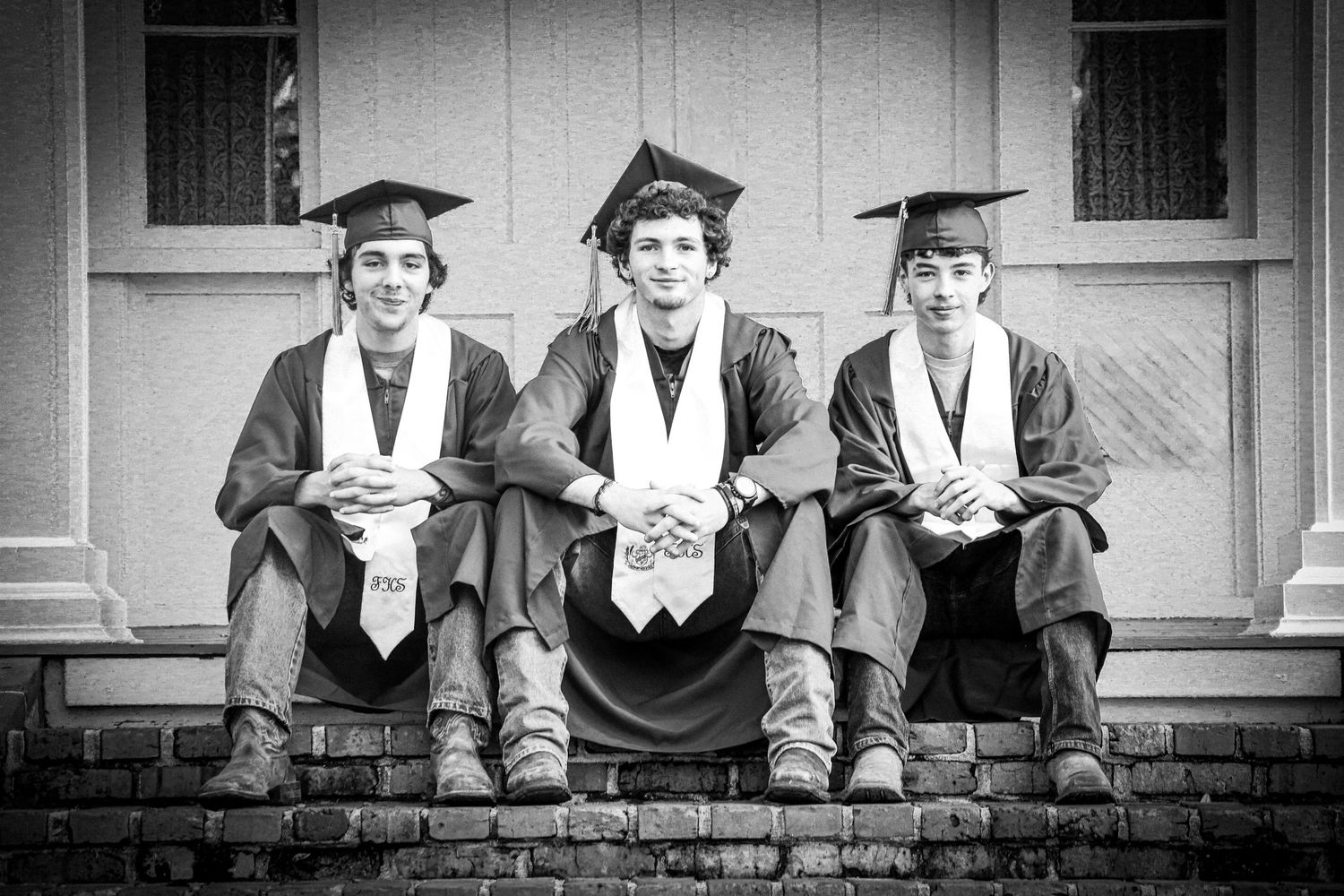 Three young male graduates in caps and gowns sit together on brick steps in a classic black and white graduation photo. seniors