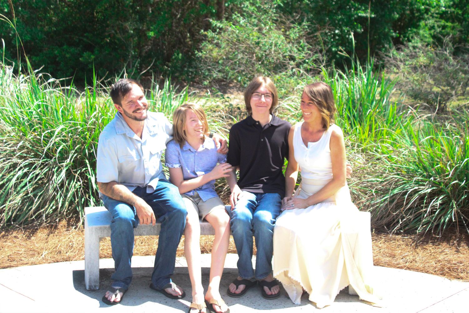 Family of four smiling and sitting together on a bench outdoors surrounded by lush green plants on a sunny day.