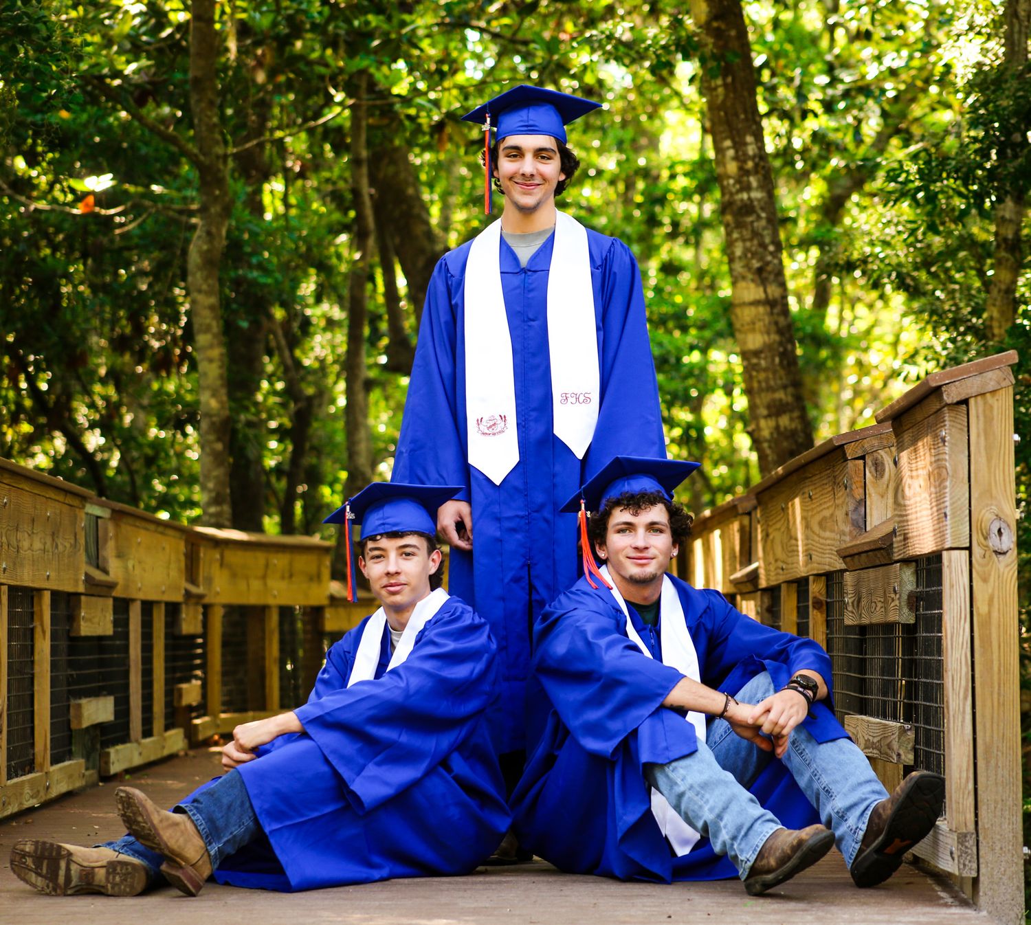 Three young men in blue graduation caps and gowns pose on a wooden bridge surrounded by lush green trees. seniors