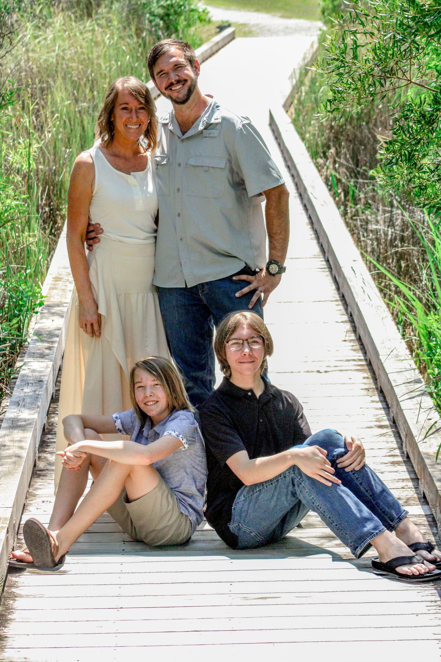Family of four posing on a wooden boardwalk surrounded by lush greenery on a sunny day.