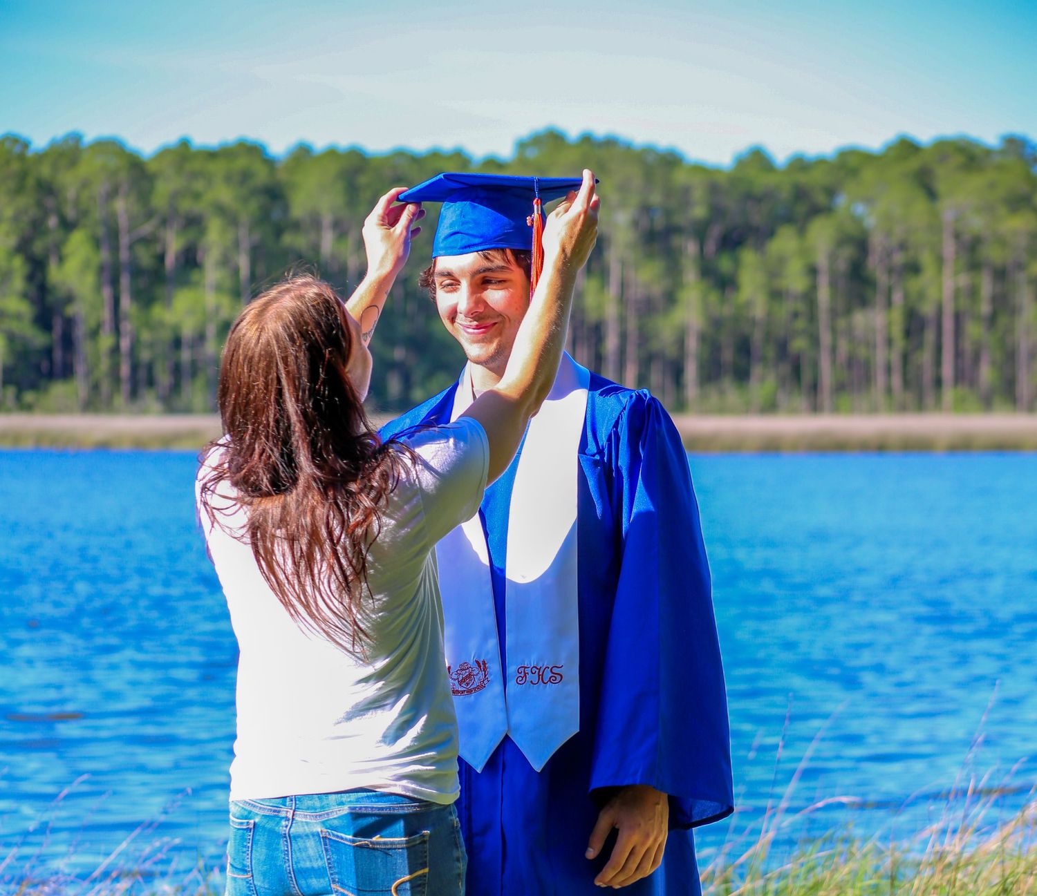Woman adjusts blue graduation cap on smiling young man in blue gown near scenic lake. mother and son