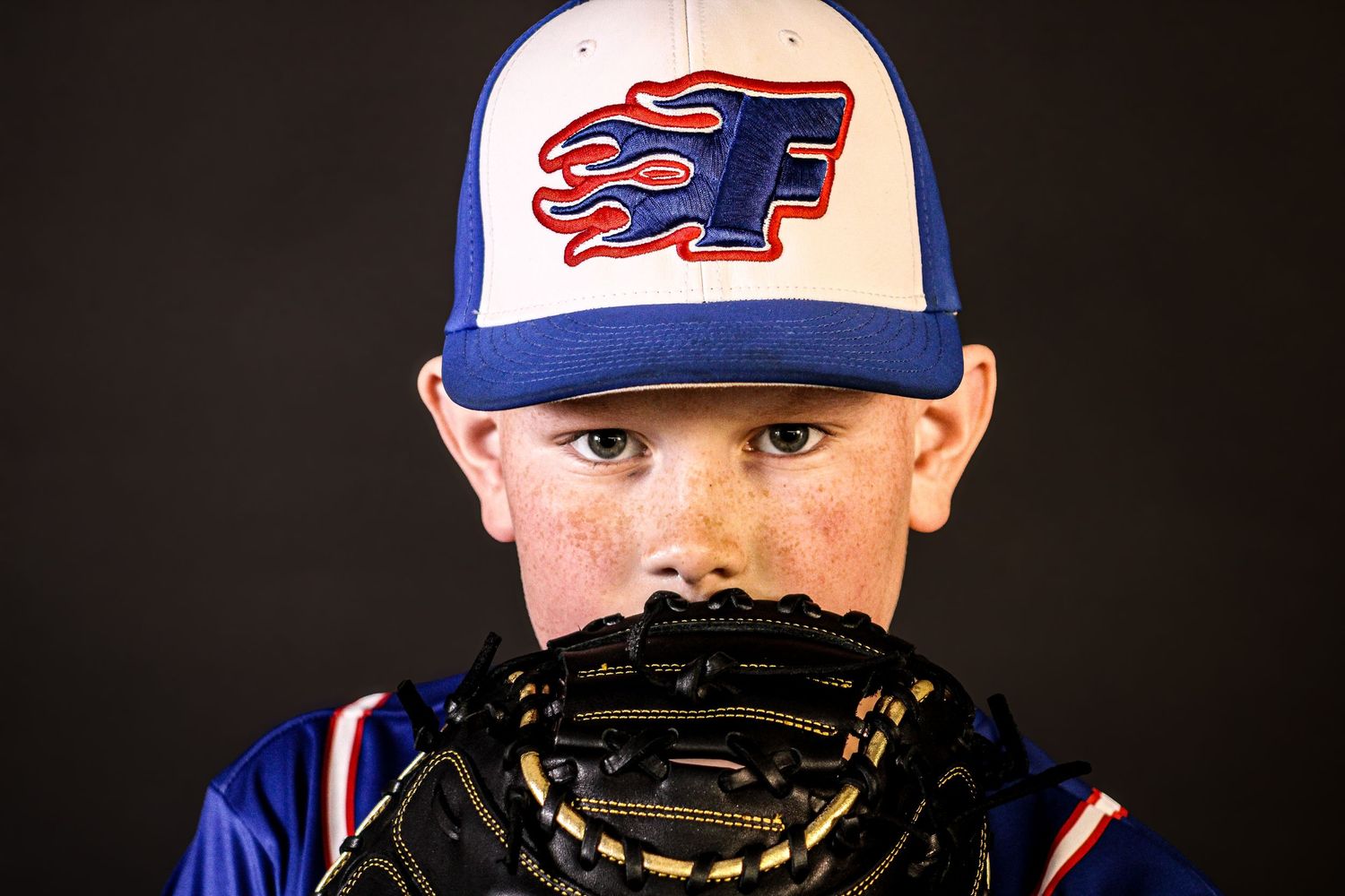 Freckled youth baseball player peers over black catcher's mitt wearing blue Flames team cap. sports