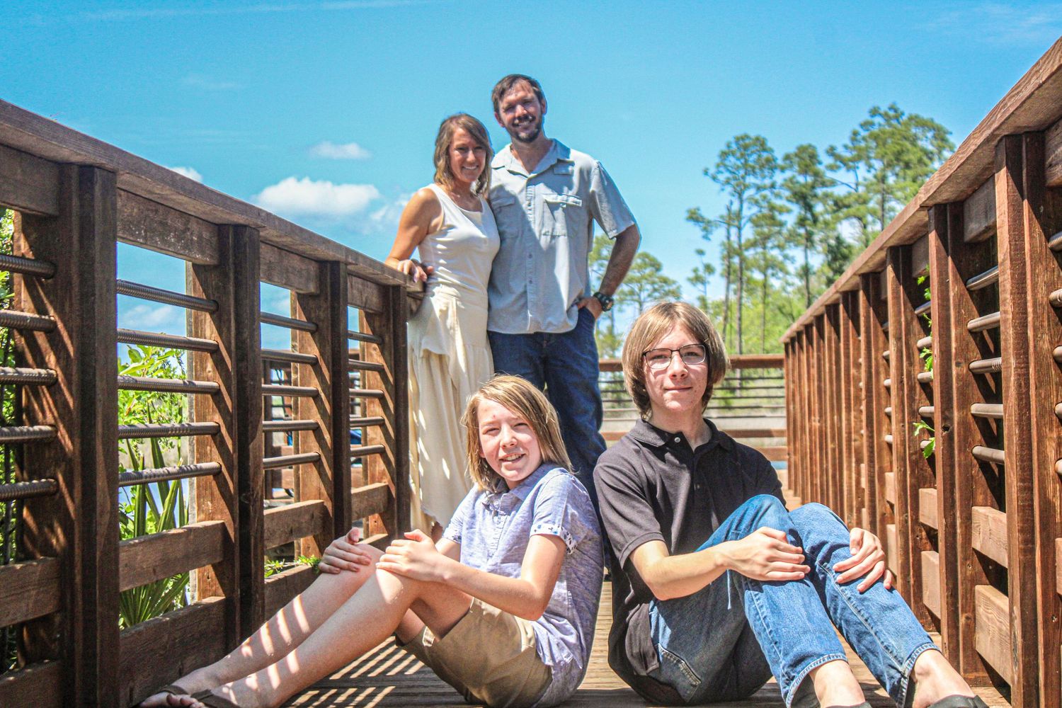 Family of four posing on a wooden bridge surrounded by lush green trees on a sunny day.