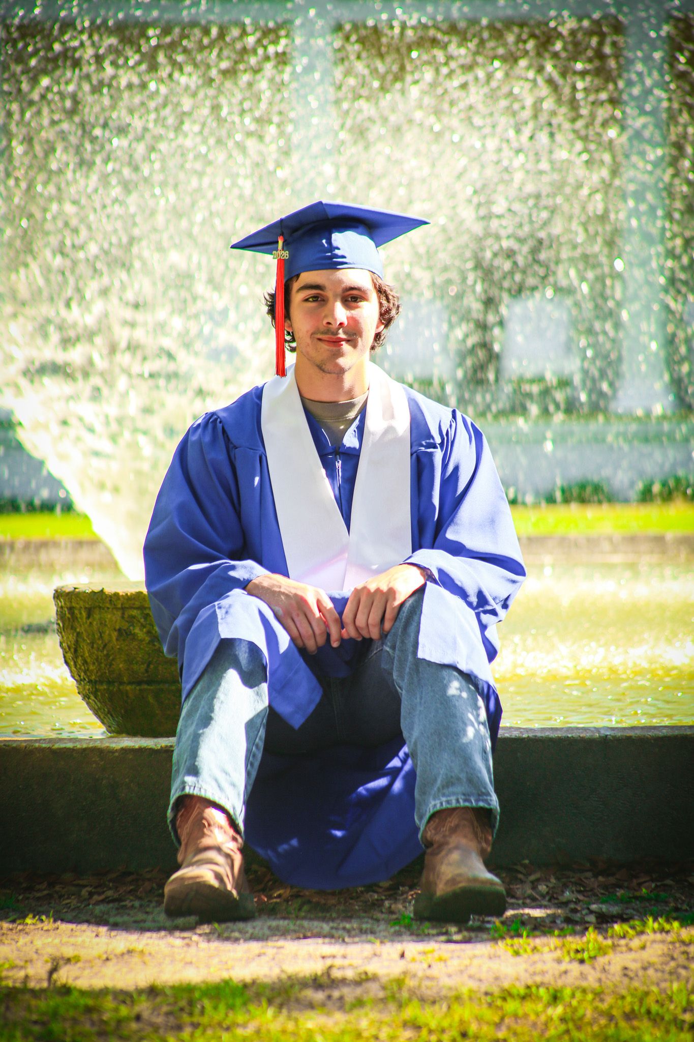 Young man in blue graduation cap and gown sits smiling by a fountain outdoors on a sunny day. senior year