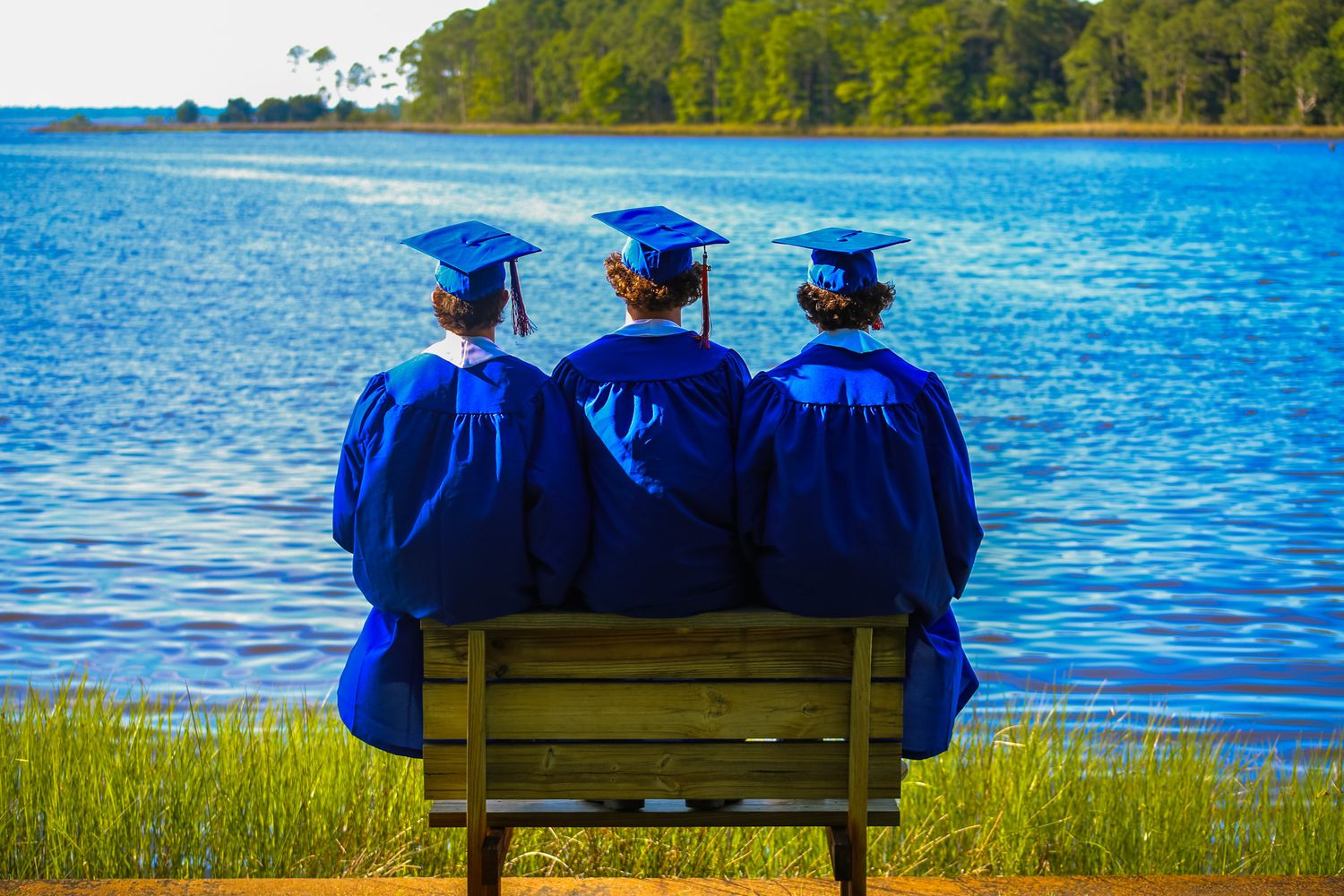 Three graduates in blue caps and gowns sit on a bench overlooking a serene lake surrounded by lush greenery. seniors