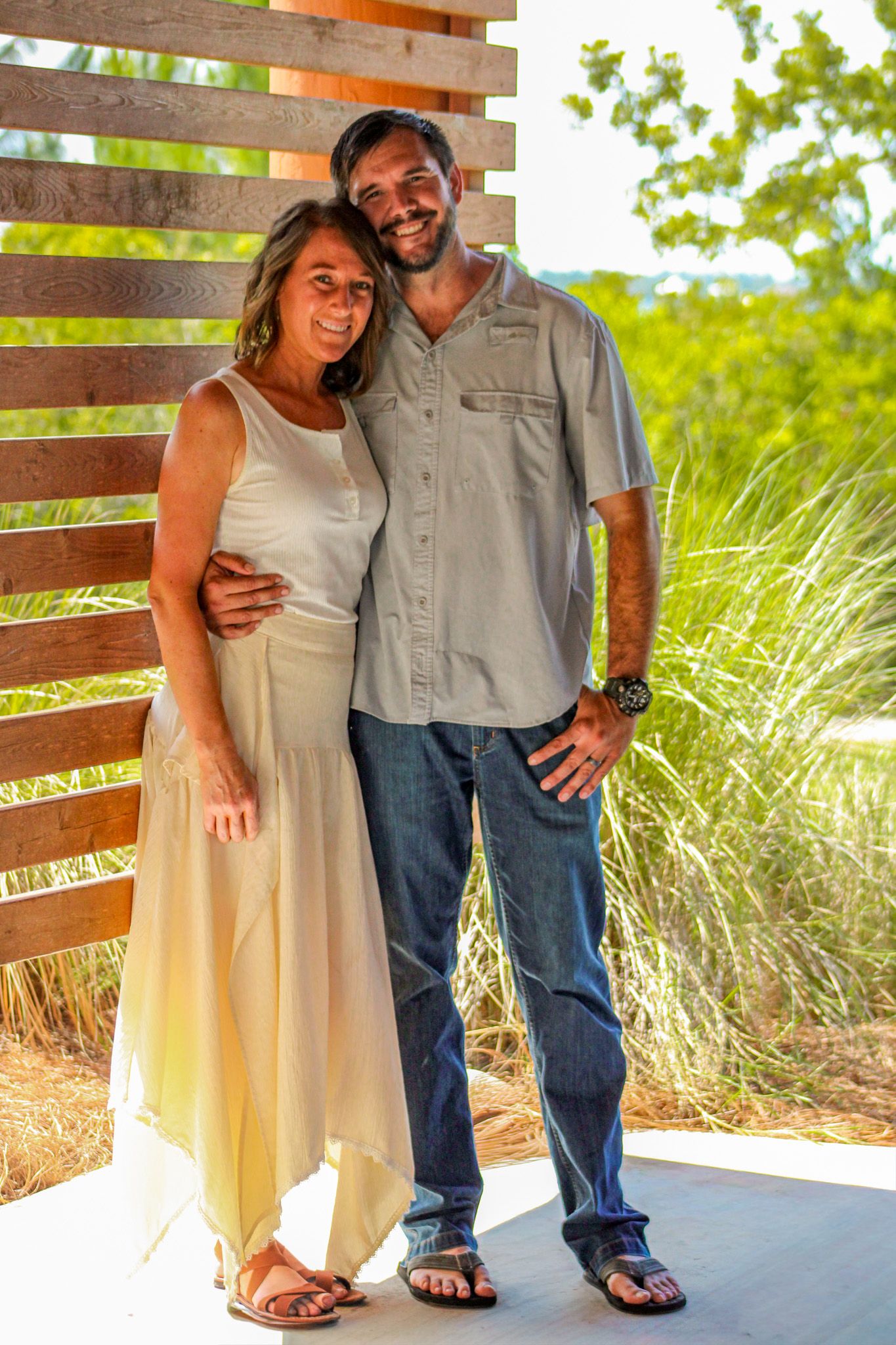 Smiling couple posing together outdoors near wooden slats and lush greenery on a sunny day.