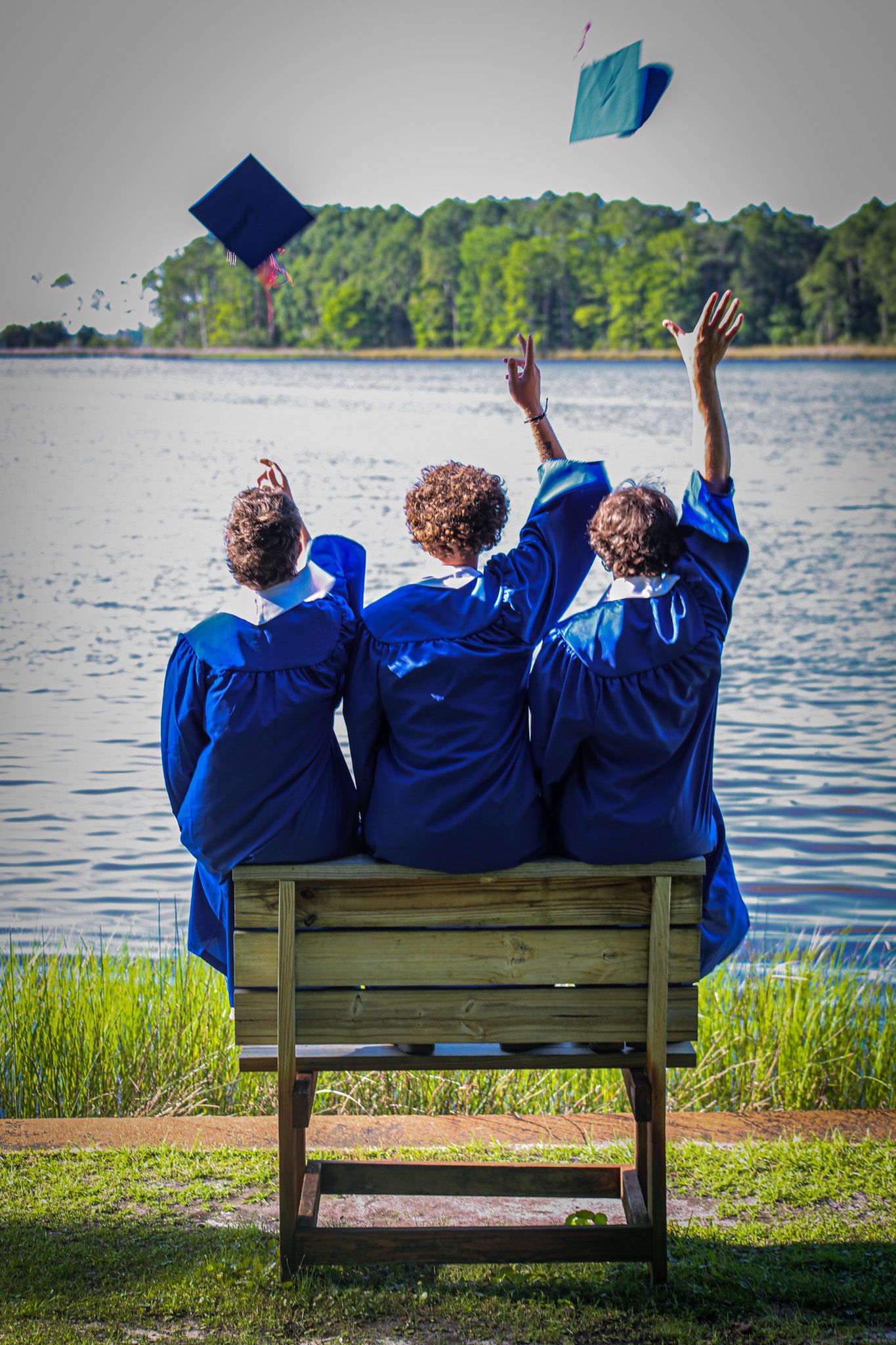 Three graduates in blue caps and gowns sit on a bench by a lake, tossing their caps into the air in celebration.