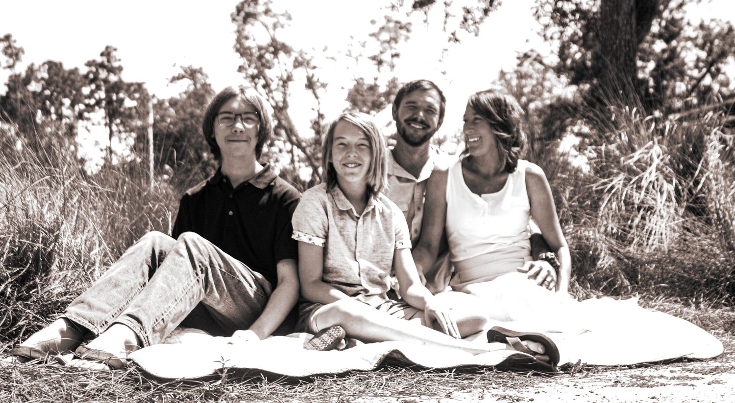 Four people sitting together on a blanket outdoors in a sunny, tree-lined setting in a black and white photo.