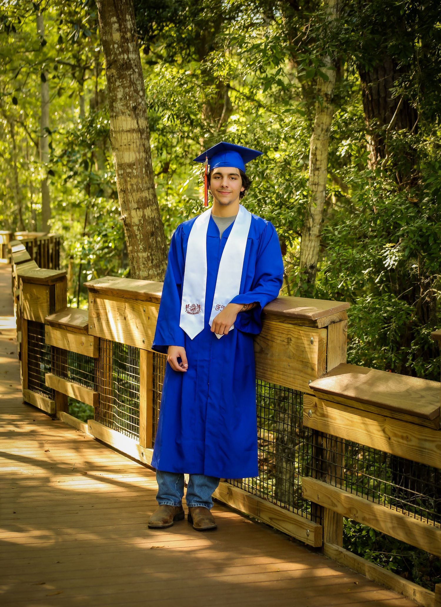 Young man in blue graduation cap and gown smiling on a wooden bridge surrounded by lush green trees. senior