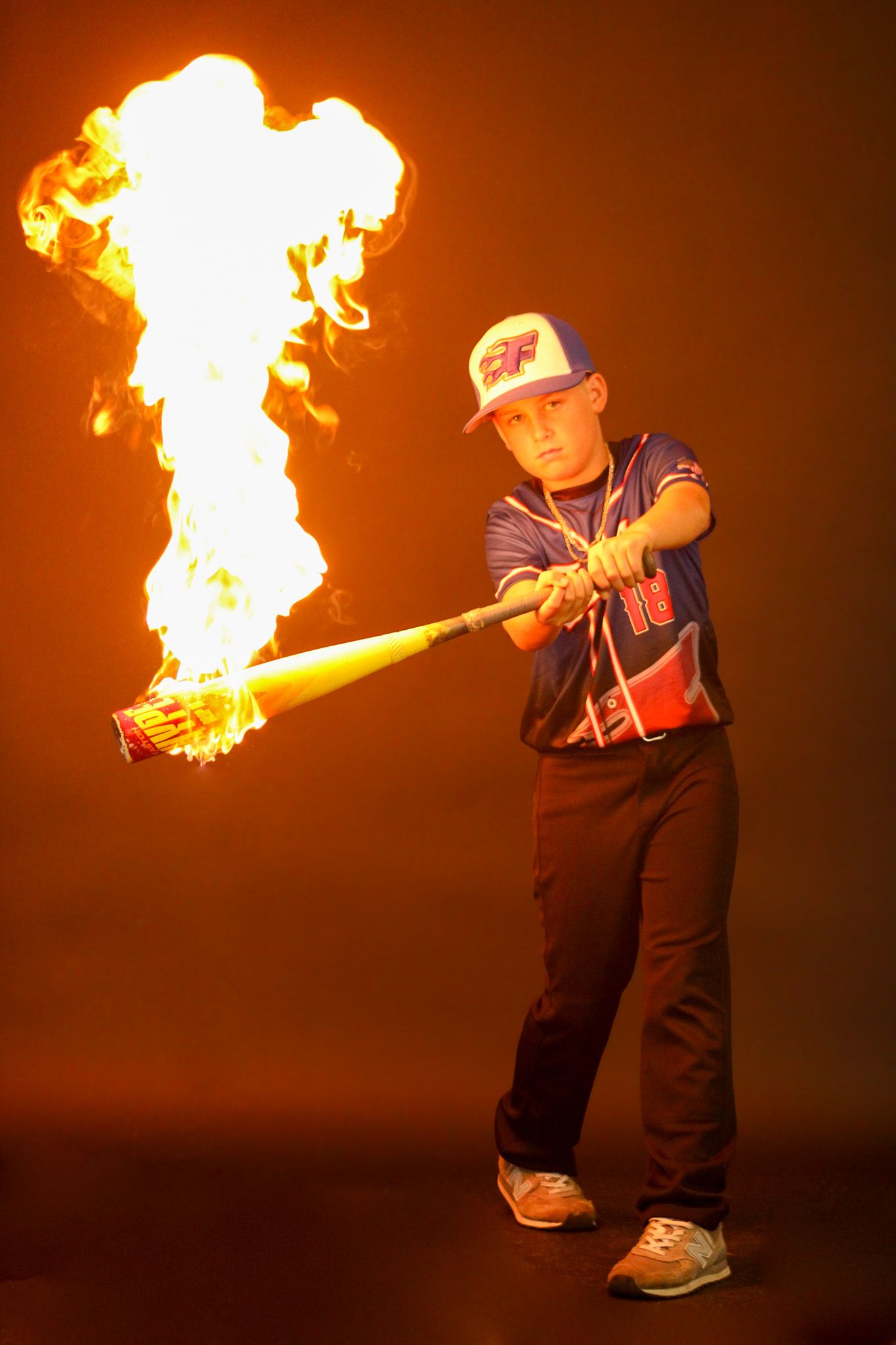 Young baseball player in uniform swinging a flaming bat with dramatic fire effect against orange background.