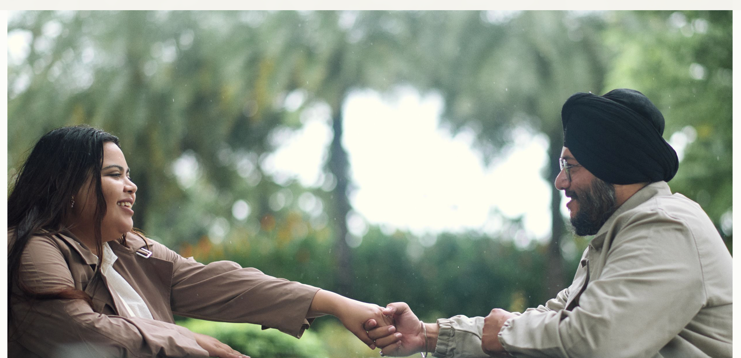 A Sikh couple holds hands and smiles at each other while sitting outdoors surrounded by lush green trees.