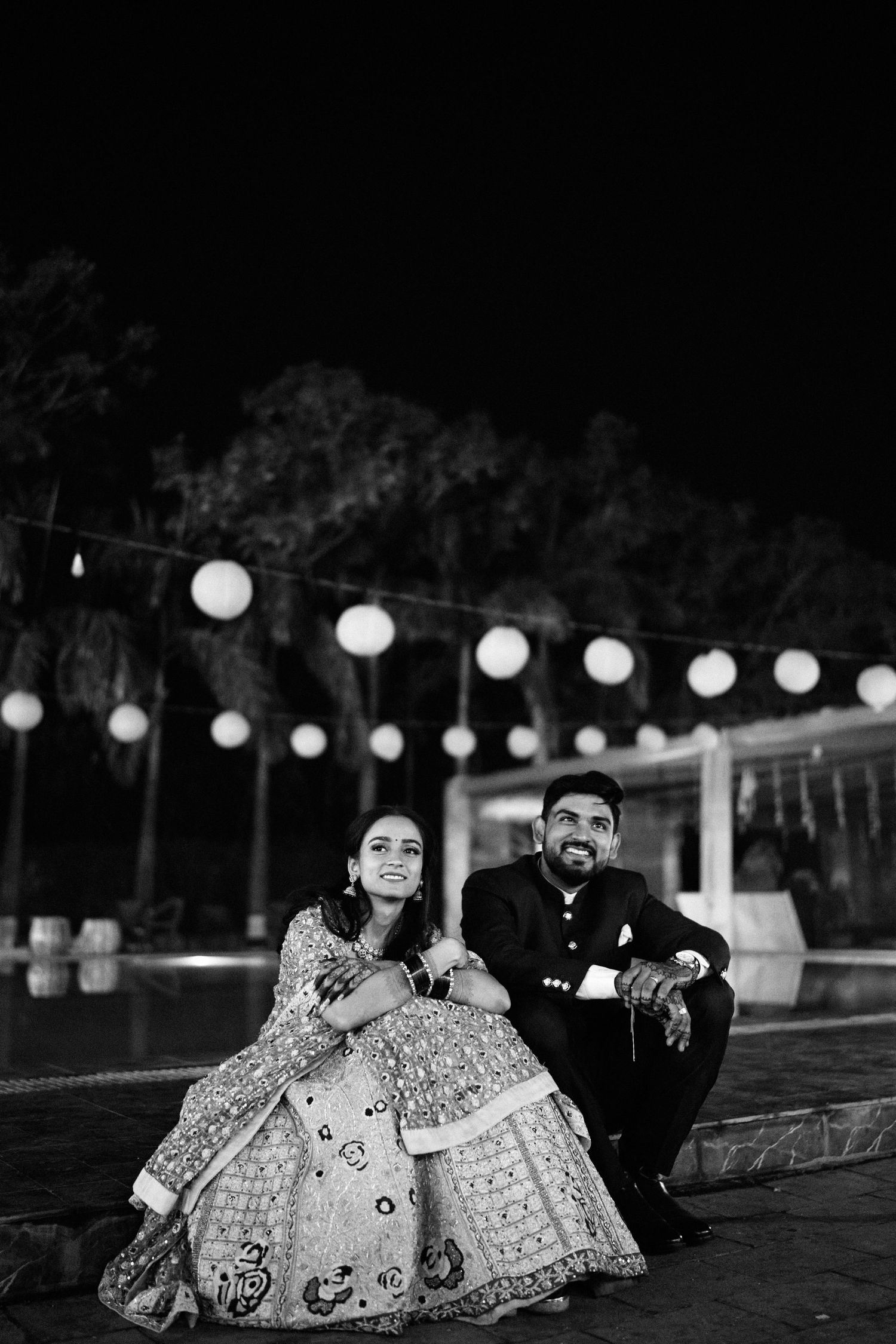 Black and white photo of an Indian couple in traditional attire sitting outdoors at night with festive string lights.