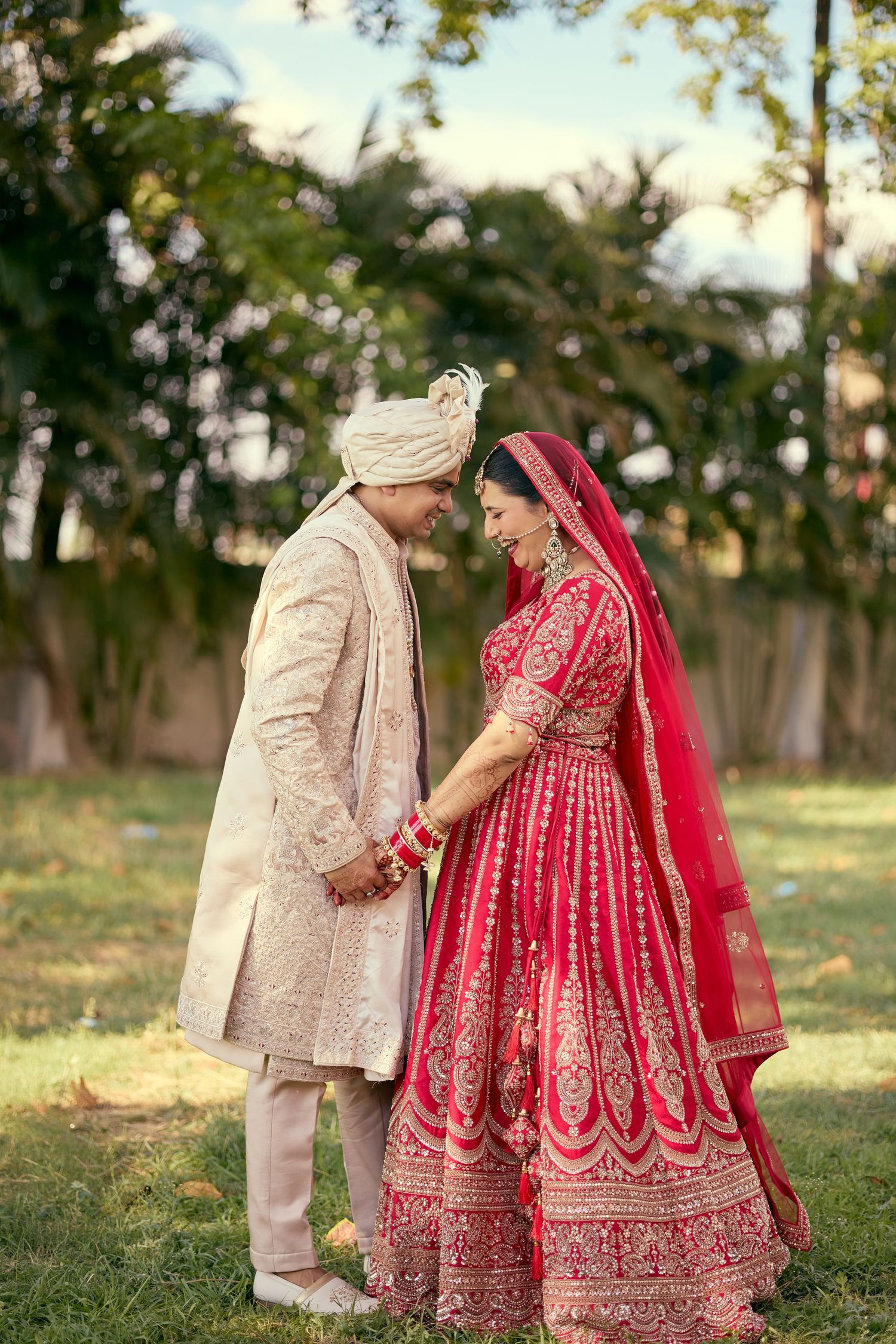 Indian bride and groom in traditional red and gold wedding attire holding hands outdoors.