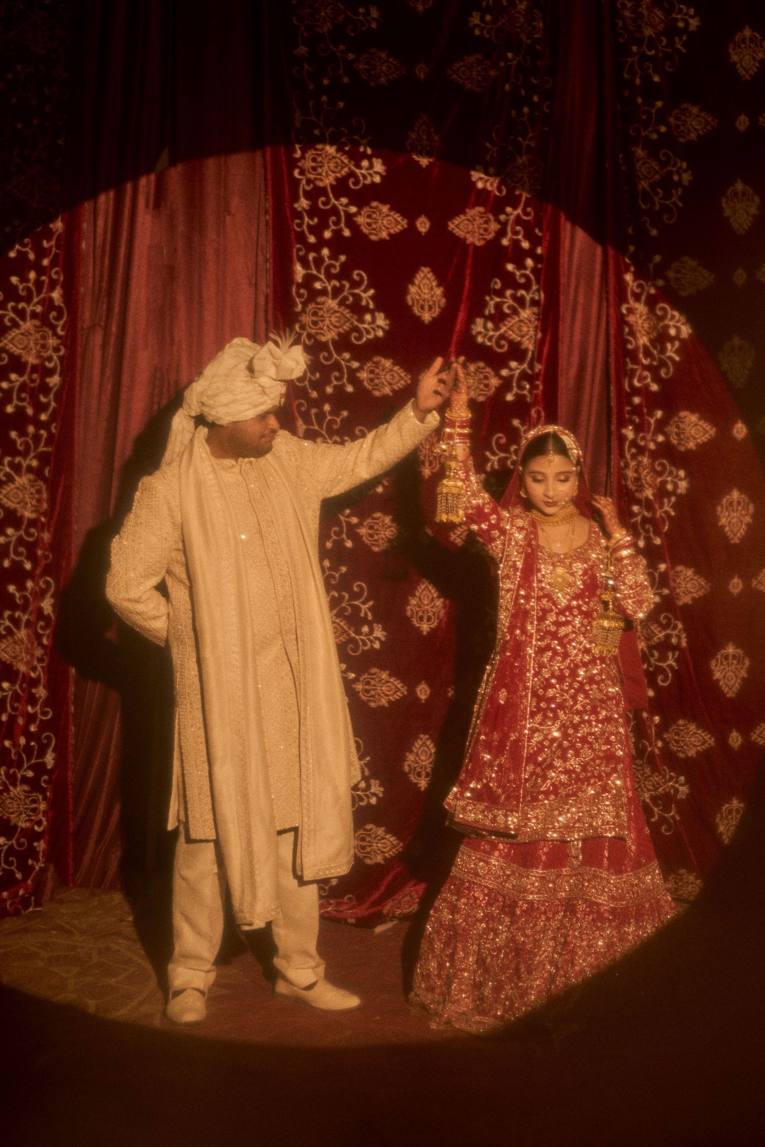 Indian bride in red lehenga and groom in cream sherwani performing wedding ritual under ornate red backdrop.
