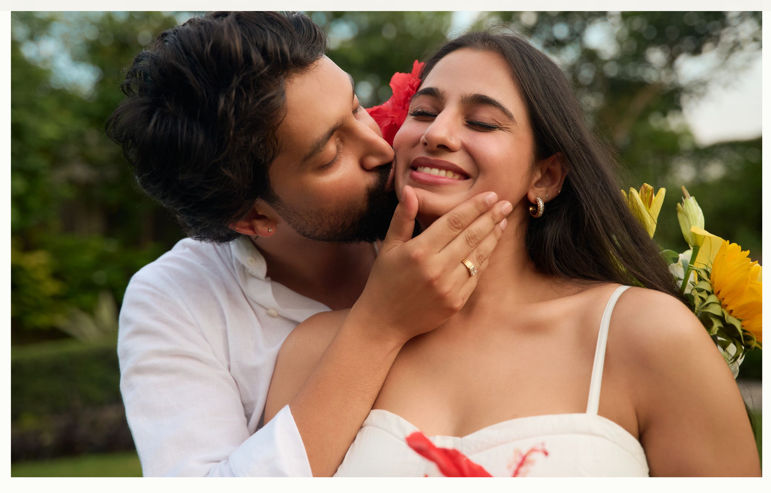 A romantic couple shares a tender kiss moment outdoors, with the man holding colorful sunflowers and a red flower.