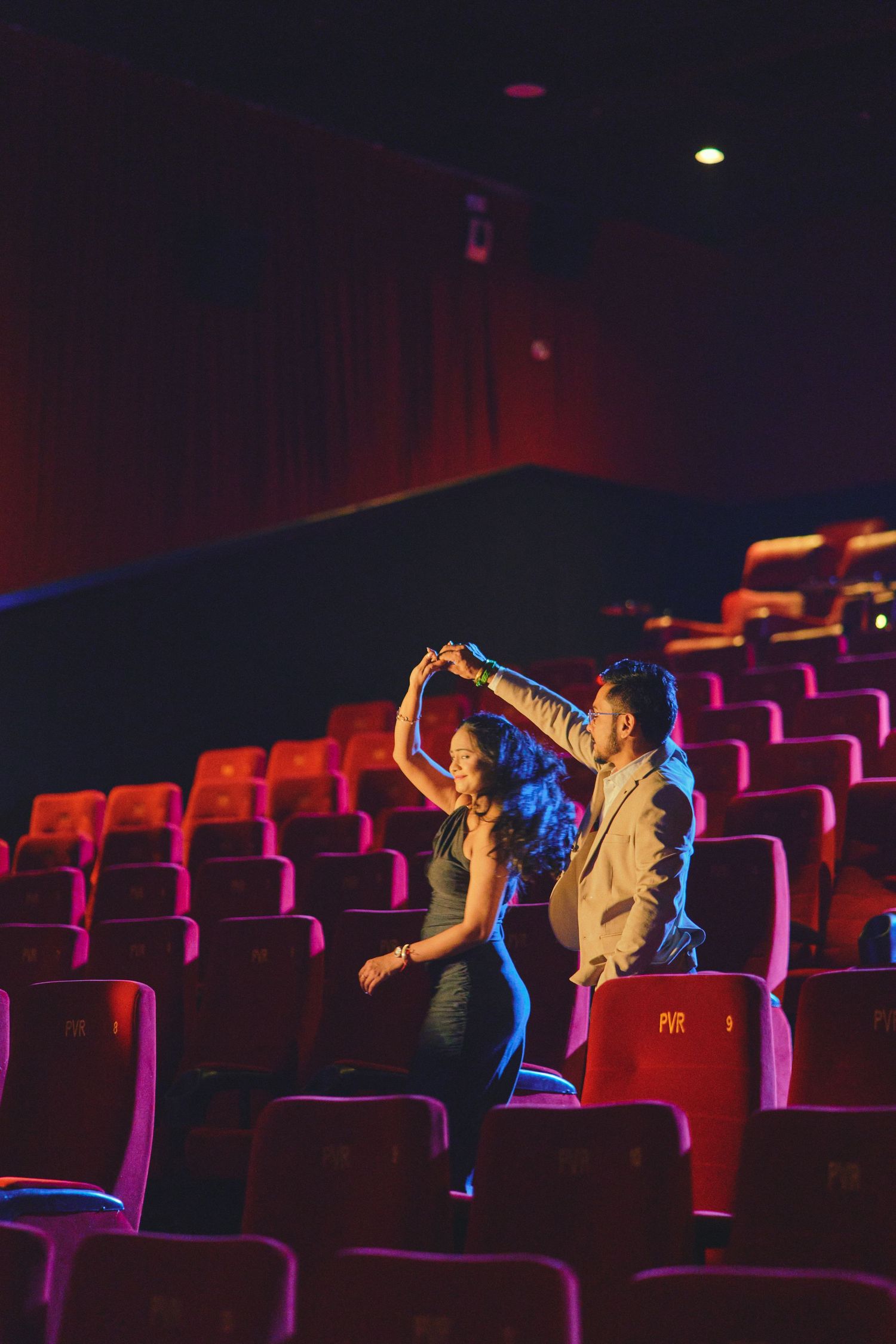 Couple dancing together in an empty red-seated movie theater, sharing a romantic moment in the dim light.